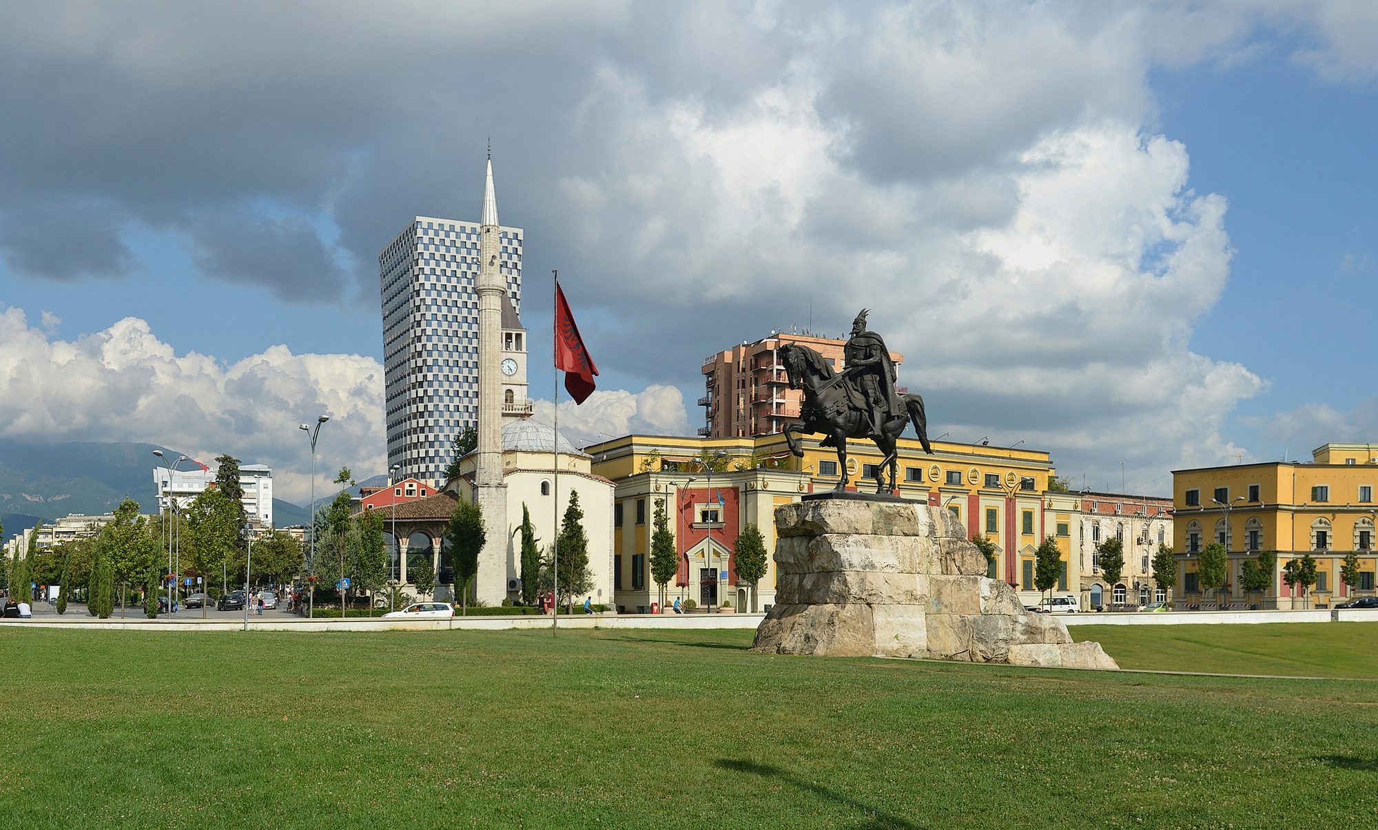 A wide open paved square with a bronze equestrian statue, minaret and clock tower behind, and mountains on the horizon.