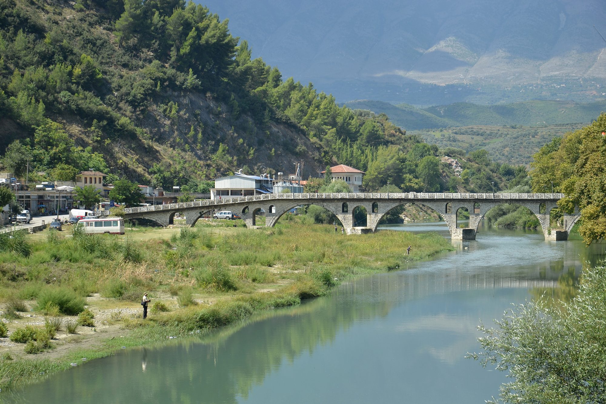 A stone arched footbridge crossing a shallow river, with wooded hills and old houses on the far bank.