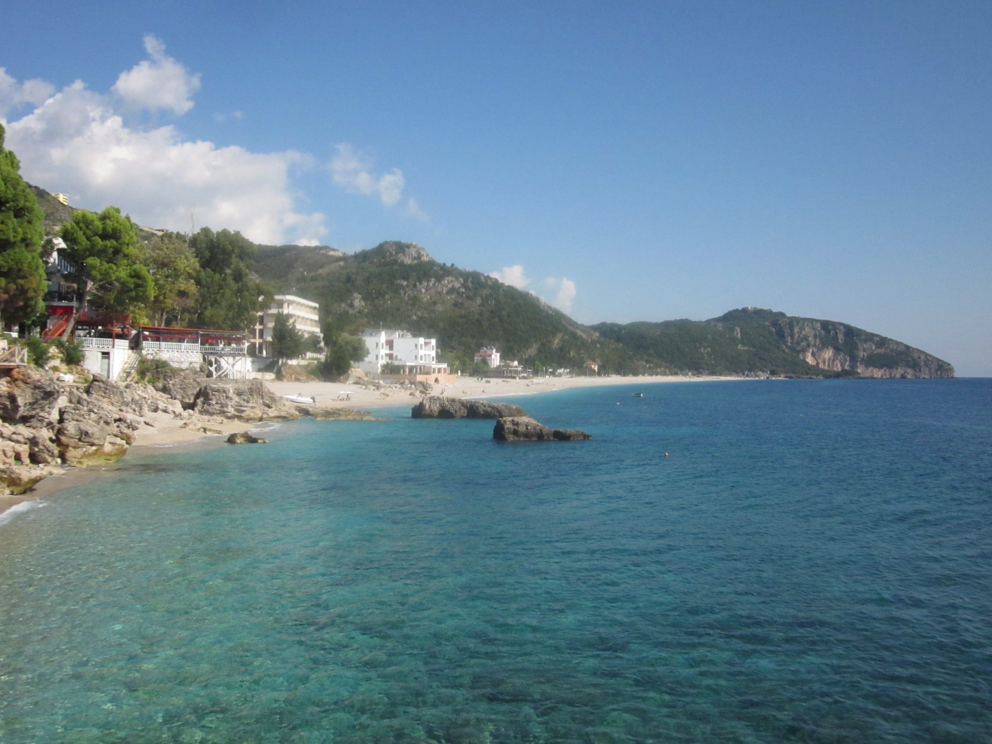 A pebble beach curving away below steep green hills, with clear turquoise water and a few sun loungers.
