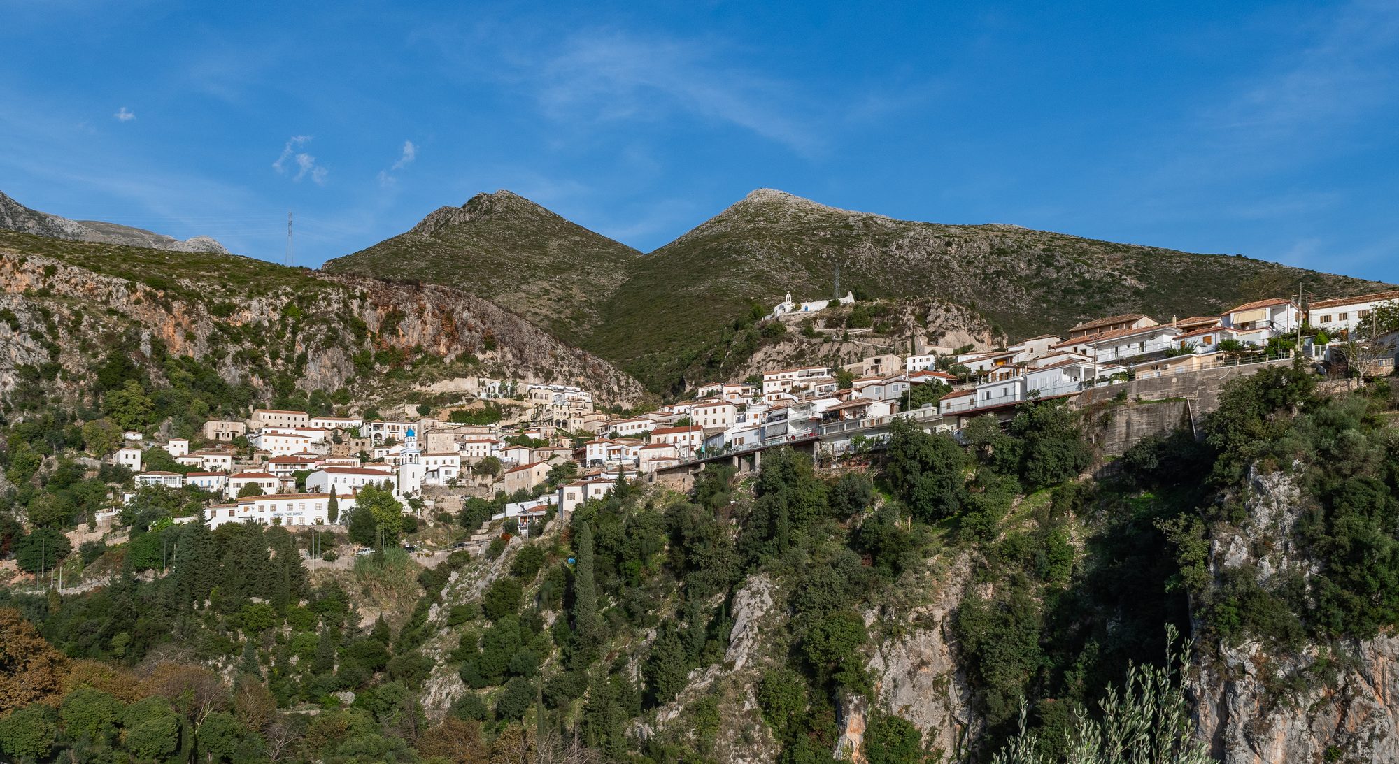 Stone houses of Dhërmi village on a mountainside, with the white monastery church of St Mary on the ridge above and the Ceraunian mountains behind.