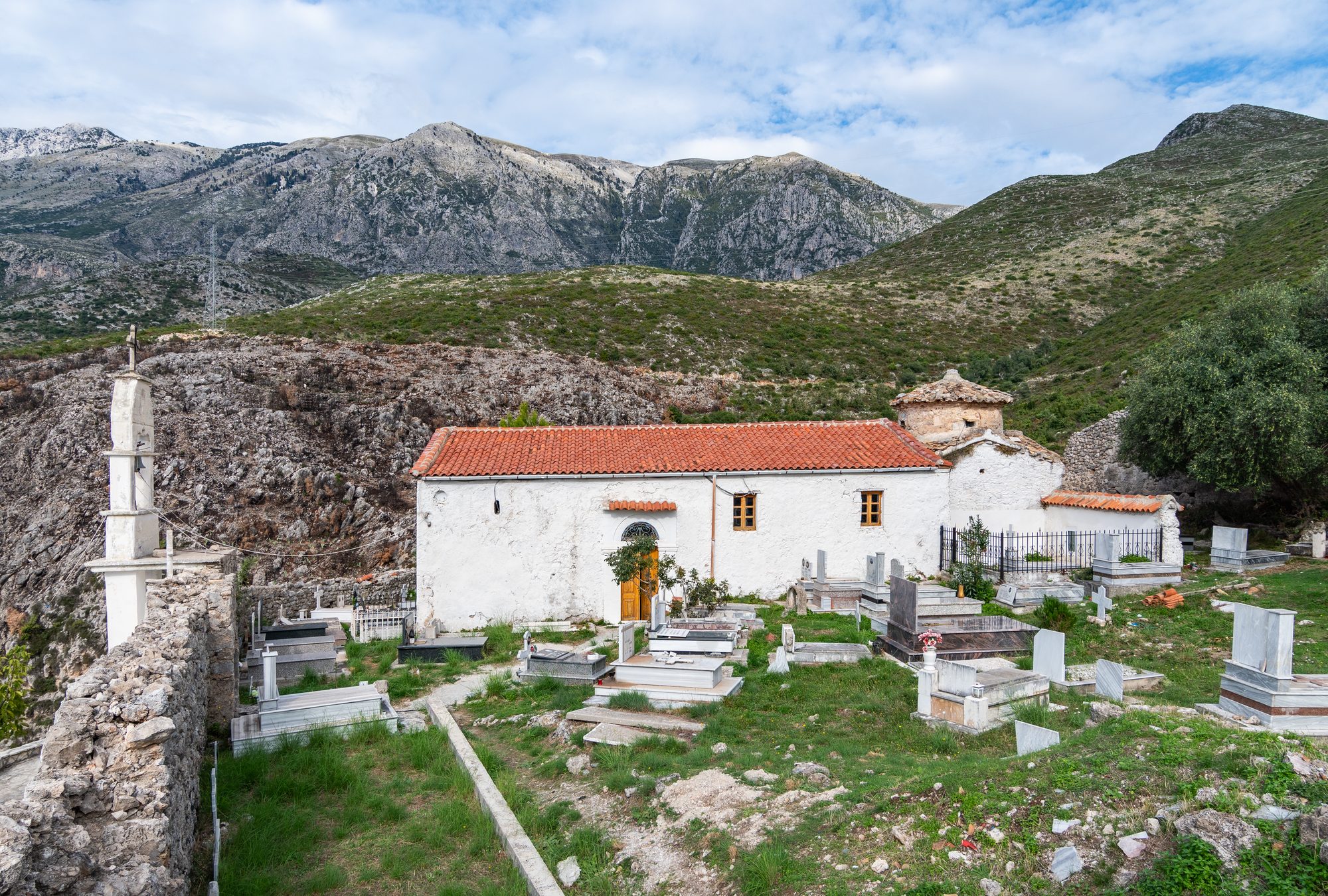 A whitewashed stone monastery church with a bell tower and cemetery, with the bare grey ridge of the Ceraunian mountains rising behind it.