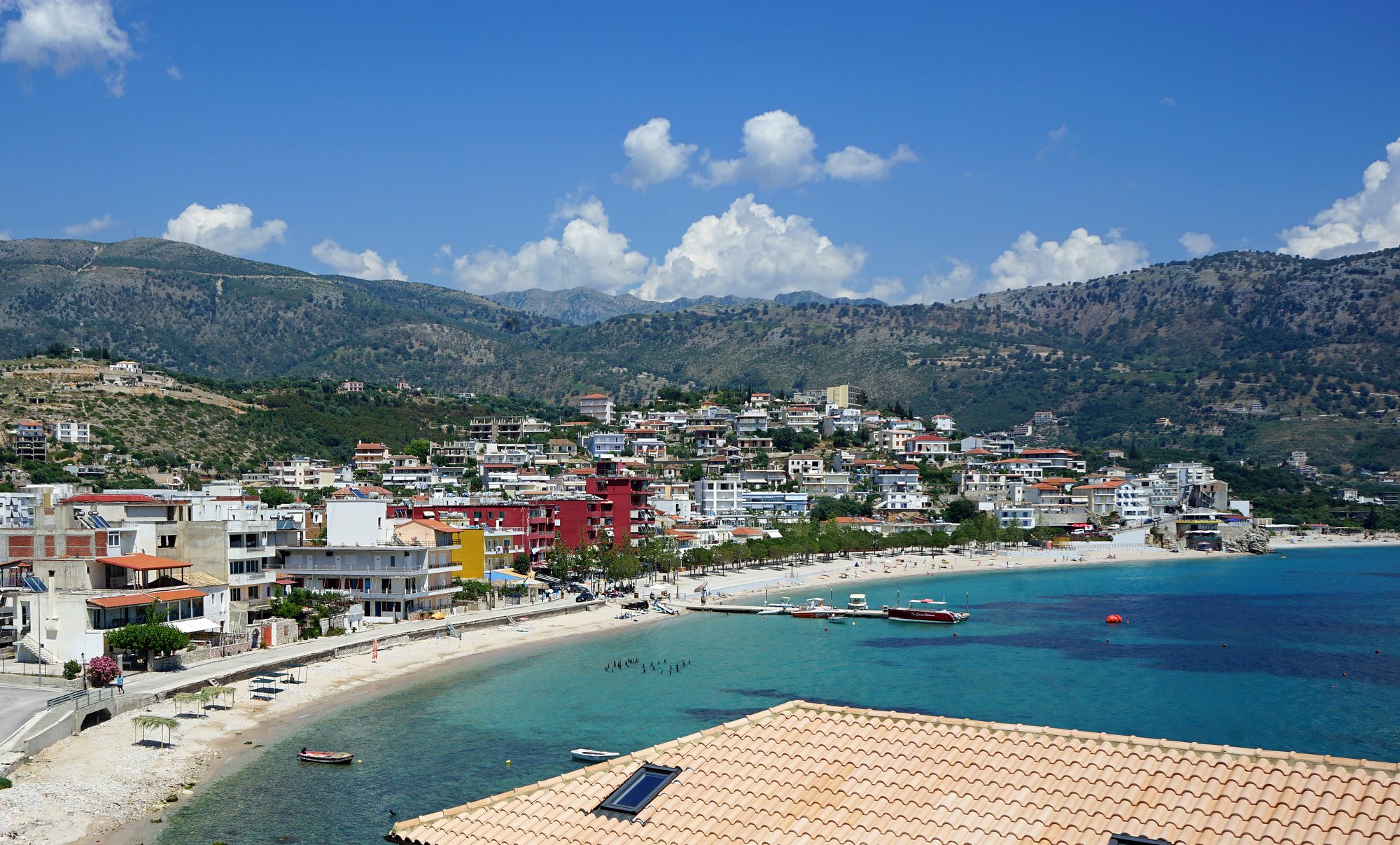A small coastal town on a hillside above a bay, with mountains rising steeply behind and clear blue sea in front.