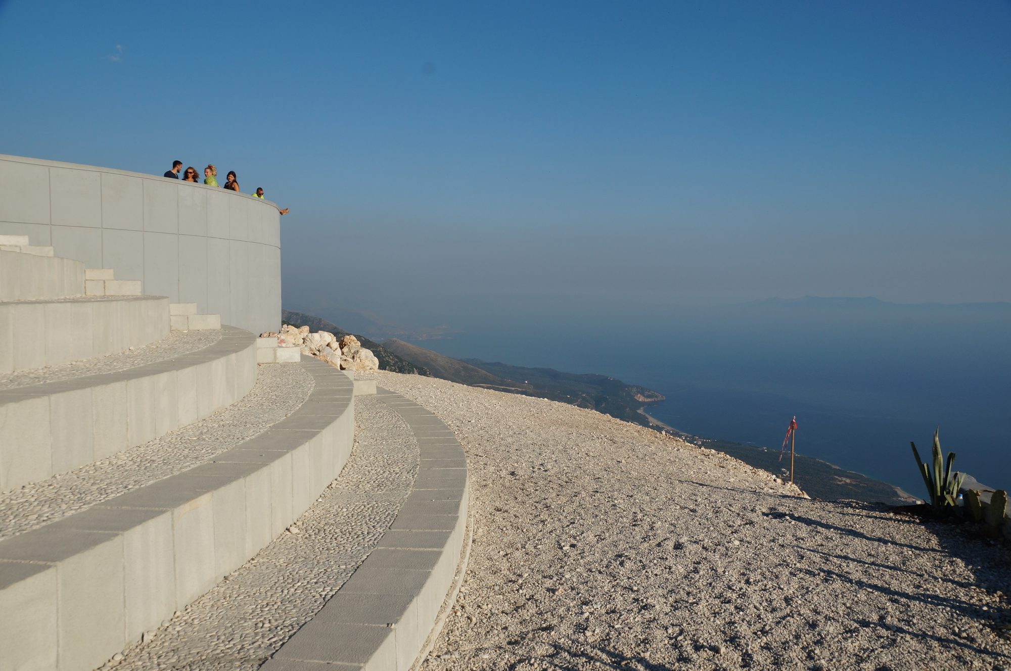 A view from a high mountain pass down onto a curving coastline, blue sea and hazy islands on the horizon.