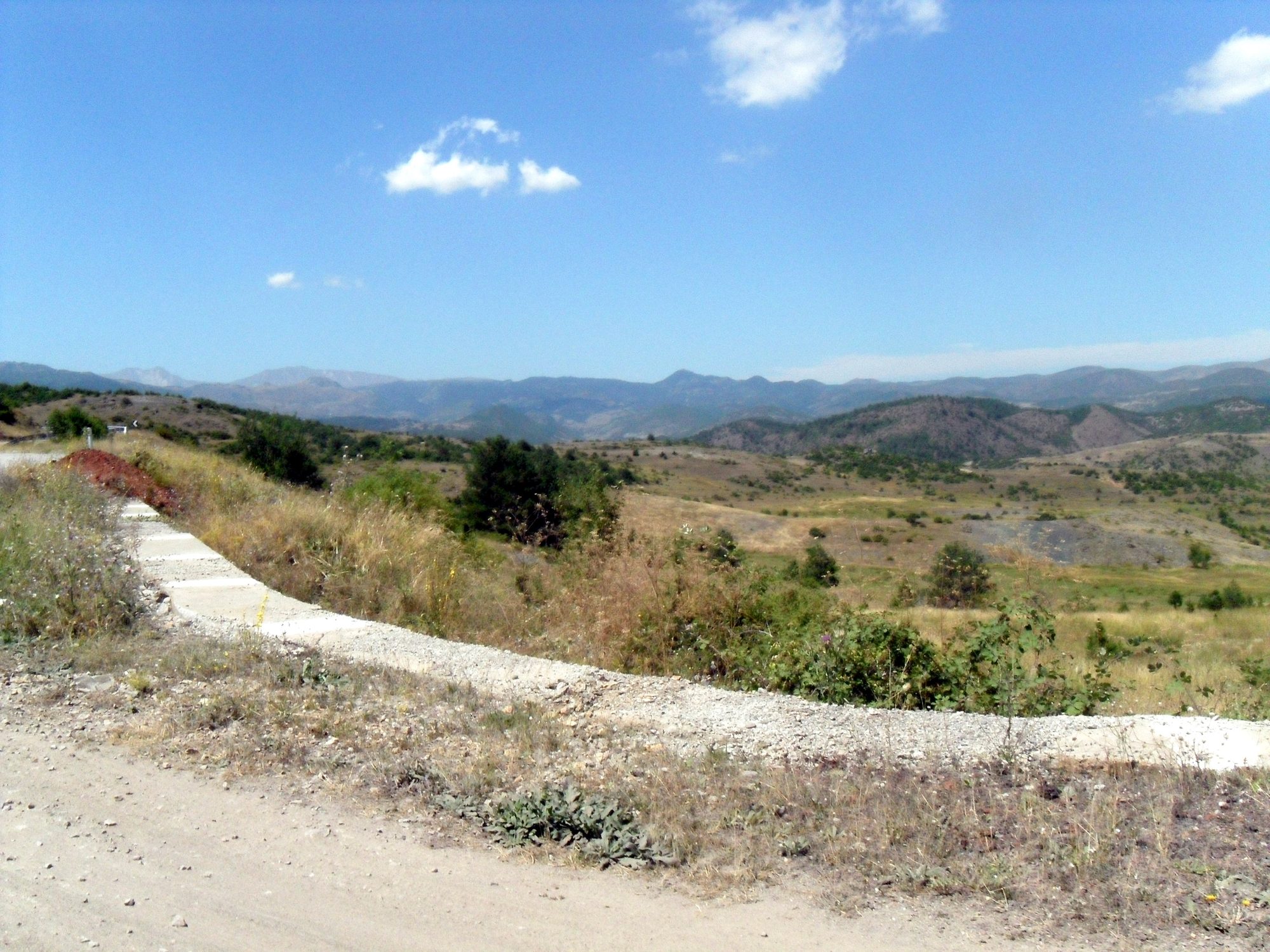 An empty asphalt road running straight through open farmland towards a long ridge of bare mountains.