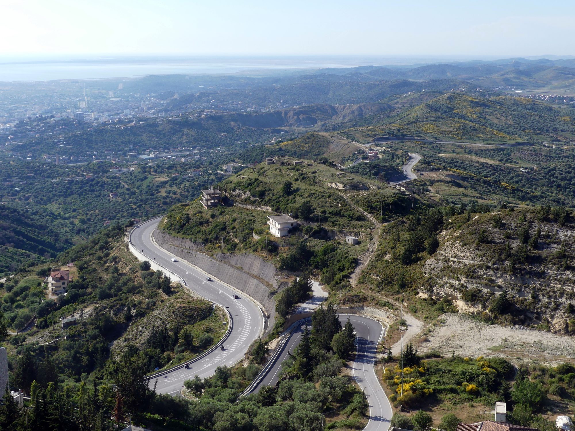 A modern two-lane road curving down through empty hills towards a distant coastal plain.