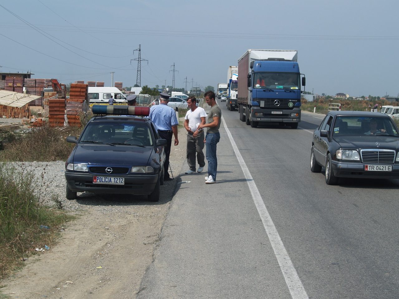 A police officer in a high-visibility vest standing beside a marked patrol car at the roadside, speaking to a stopped driver.