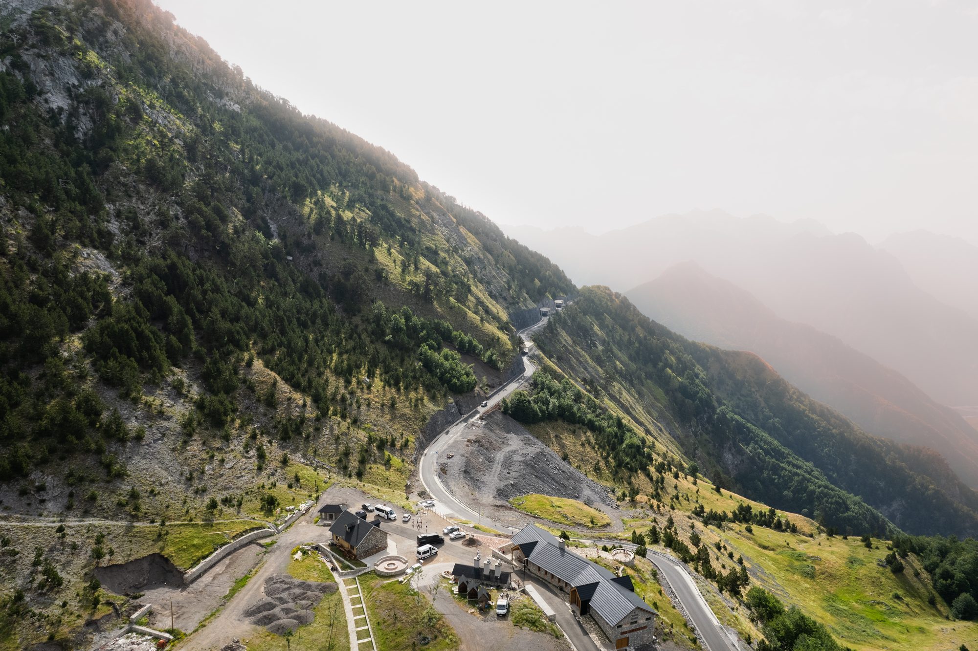 A narrow mountain road cut into a steep green slope, winding between rocky peaks under a clear sky.