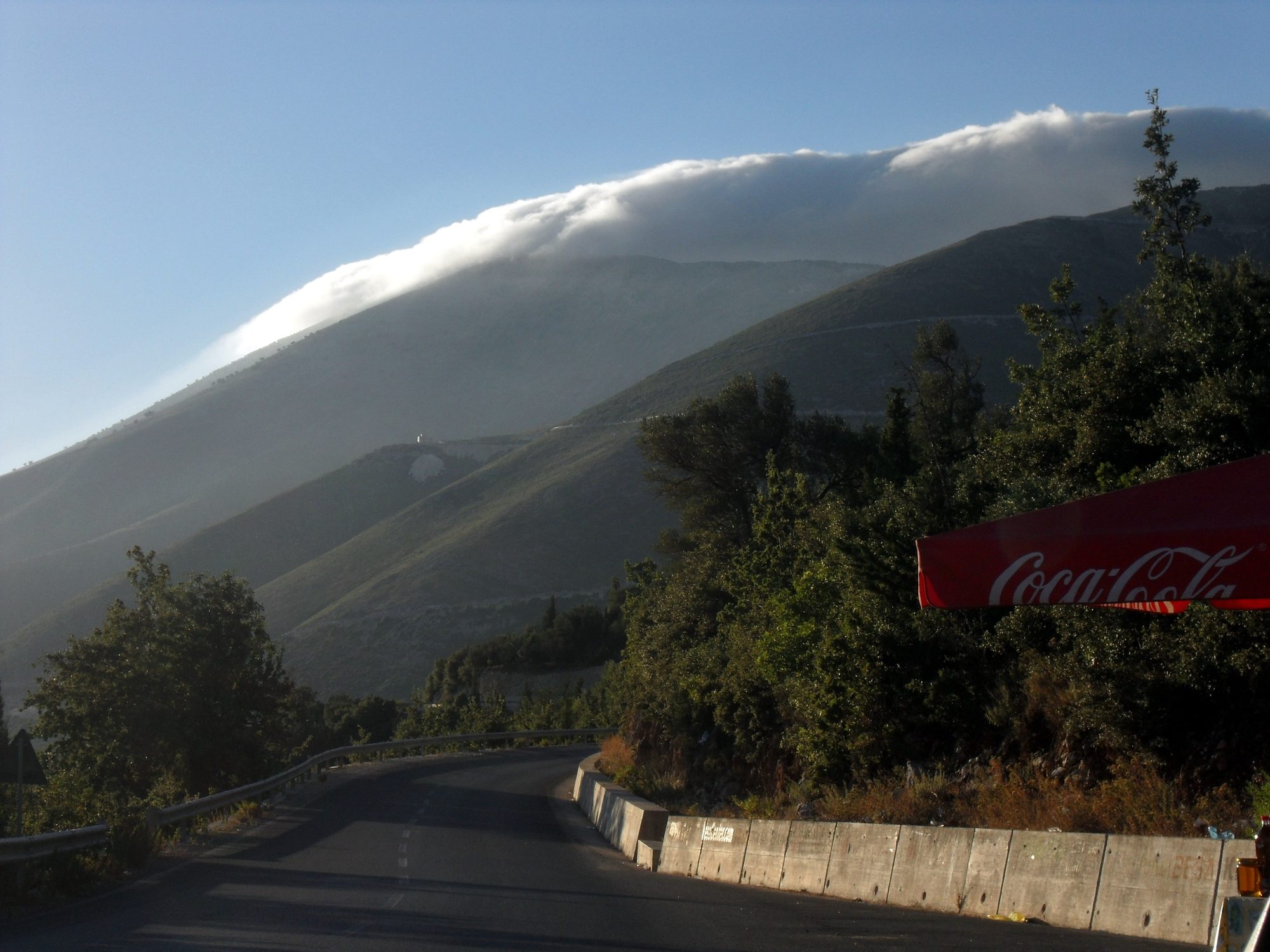 A two-lane road running along a steep coastal hillside high above a blue sea, with mountains falling away on the right.