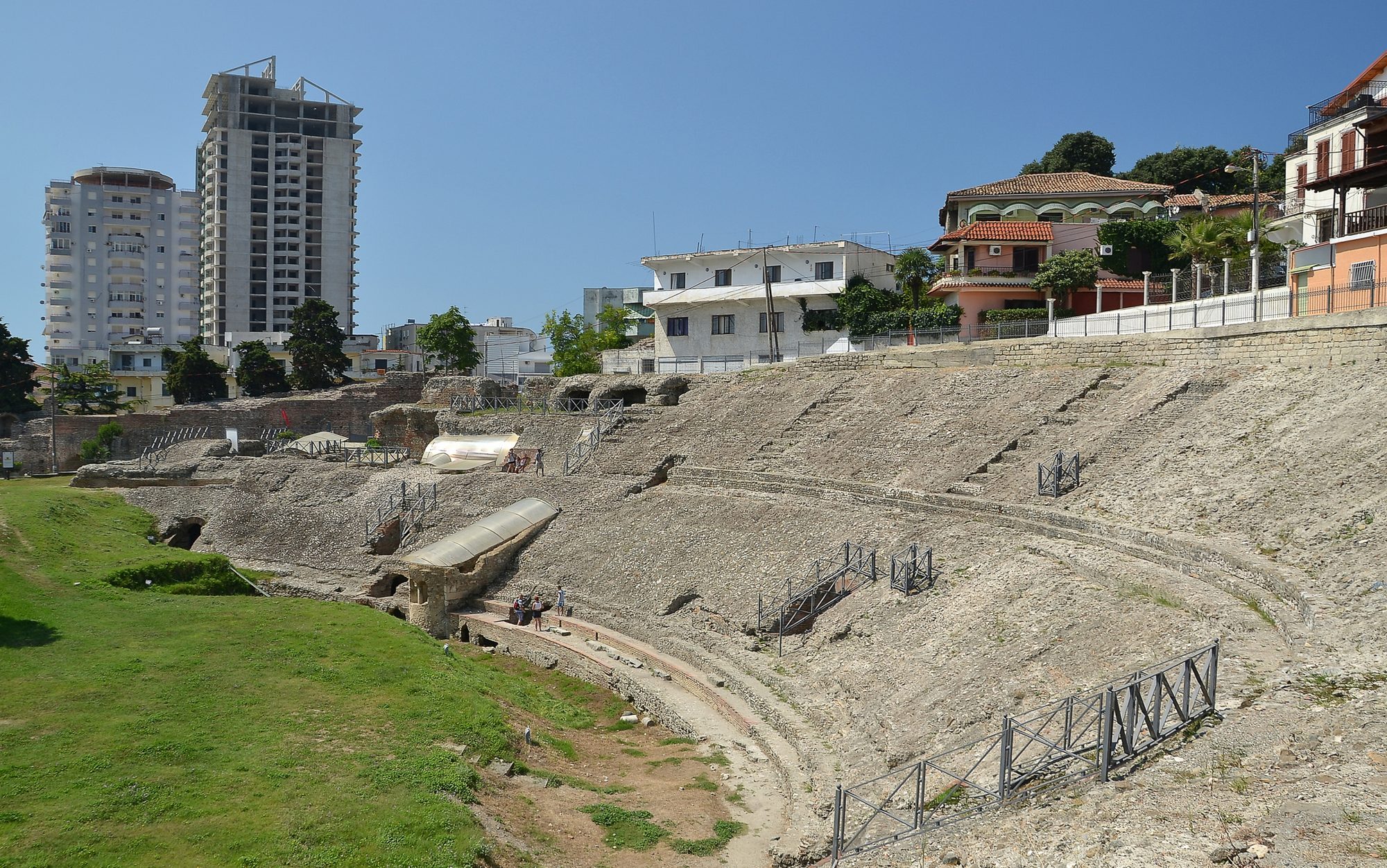 Curved stone tiers of a Roman amphitheatre partly excavated, with modern apartment buildings visible directly behind.