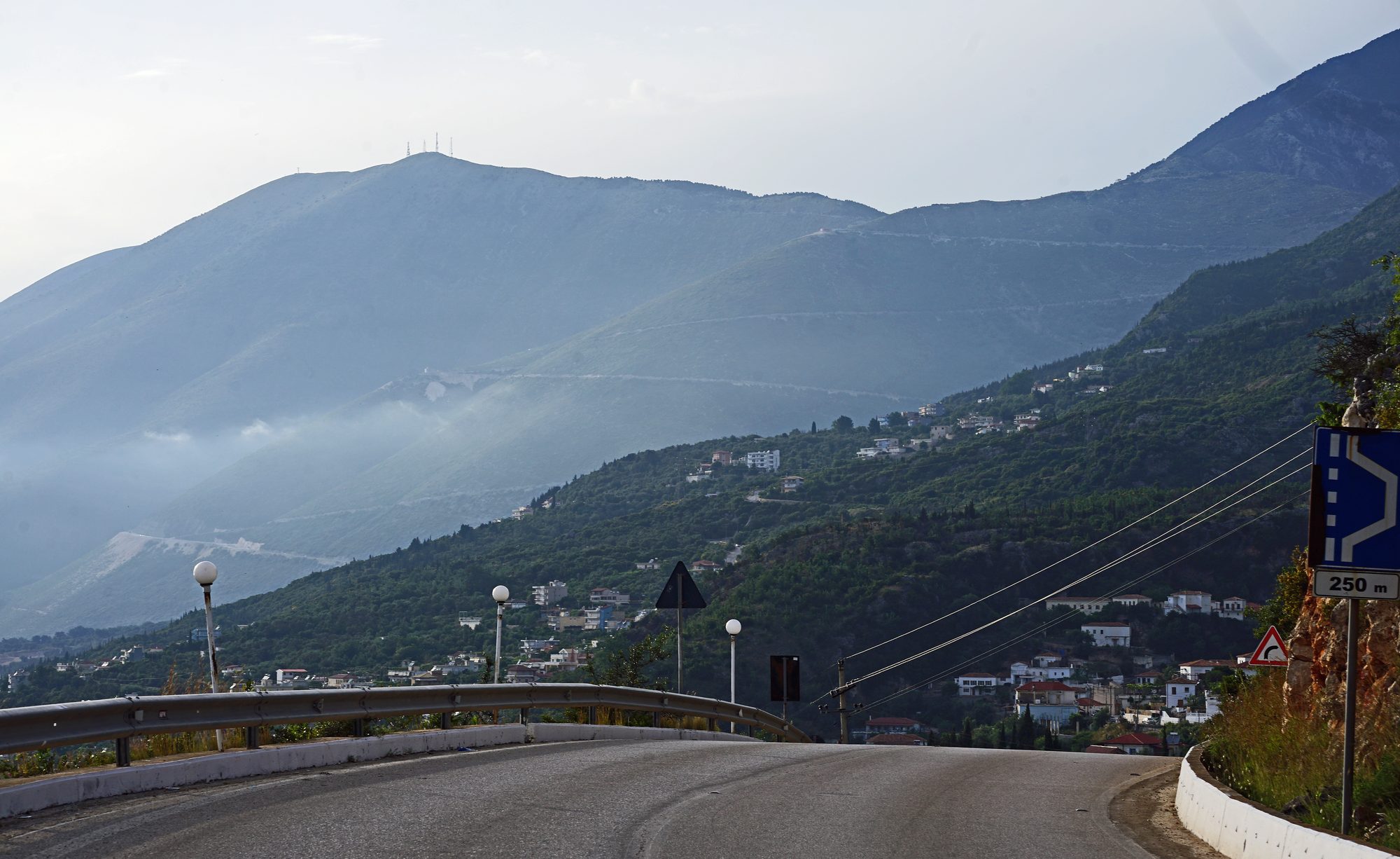 A curving coastal road descending toward Dhërmi, whose white houses are scattered across a green hillside, with the hazy ridge of the Ceraunian mountains rising behind.