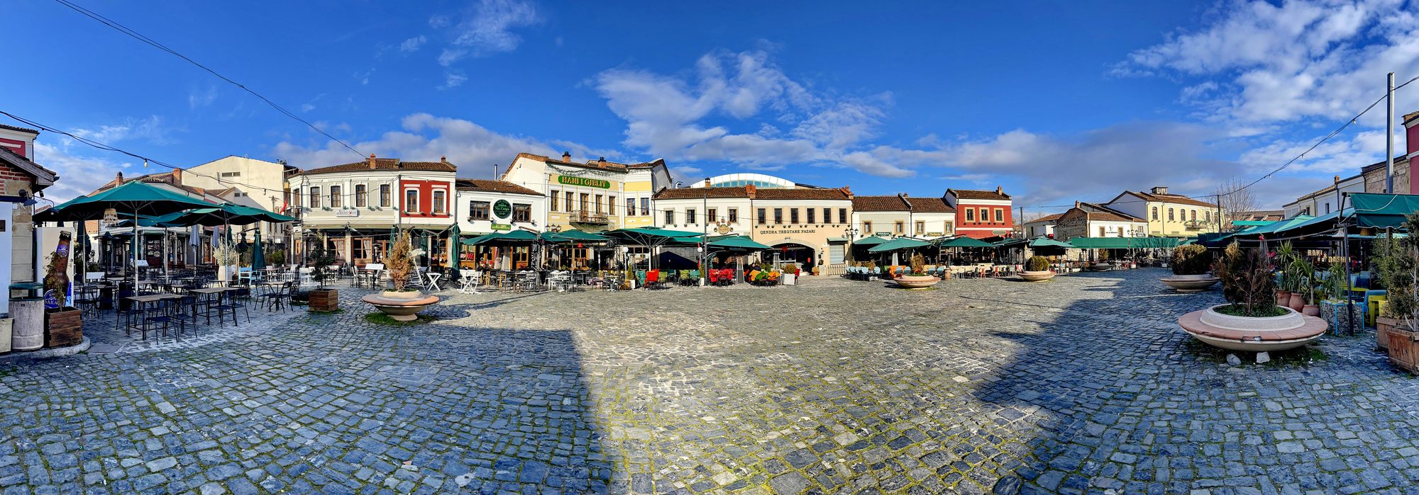 The wide cobbled square of Korçë's Old Bazaar under a bright winter sky, ringed by restored two-storey buildings with red tiled roofs and shuttered café terraces.