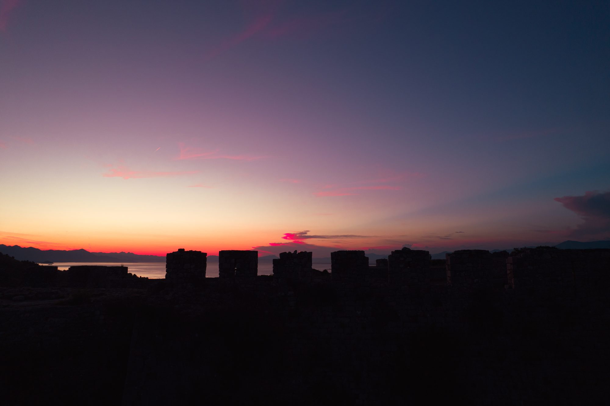The broken battlements of Rozafa Castle in silhouette against an orange and pink sunset, with the flat water of Lake Shkodra and distant mountains below.