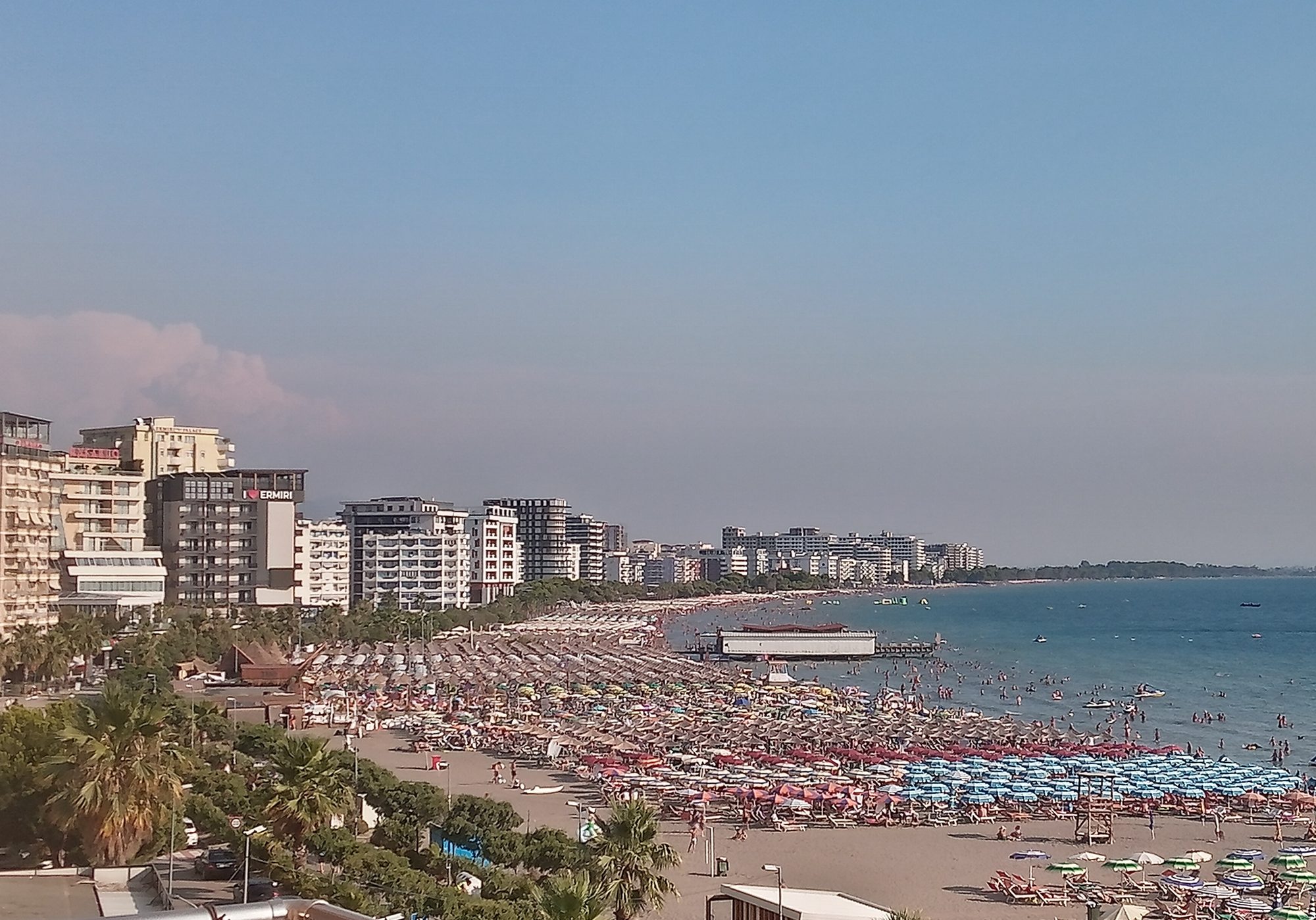 Shëngjin's beach in high summer, packed with dense rows of coloured umbrellas and sunbeds, backed by a line of tall apartment blocks and hotels along the shore.