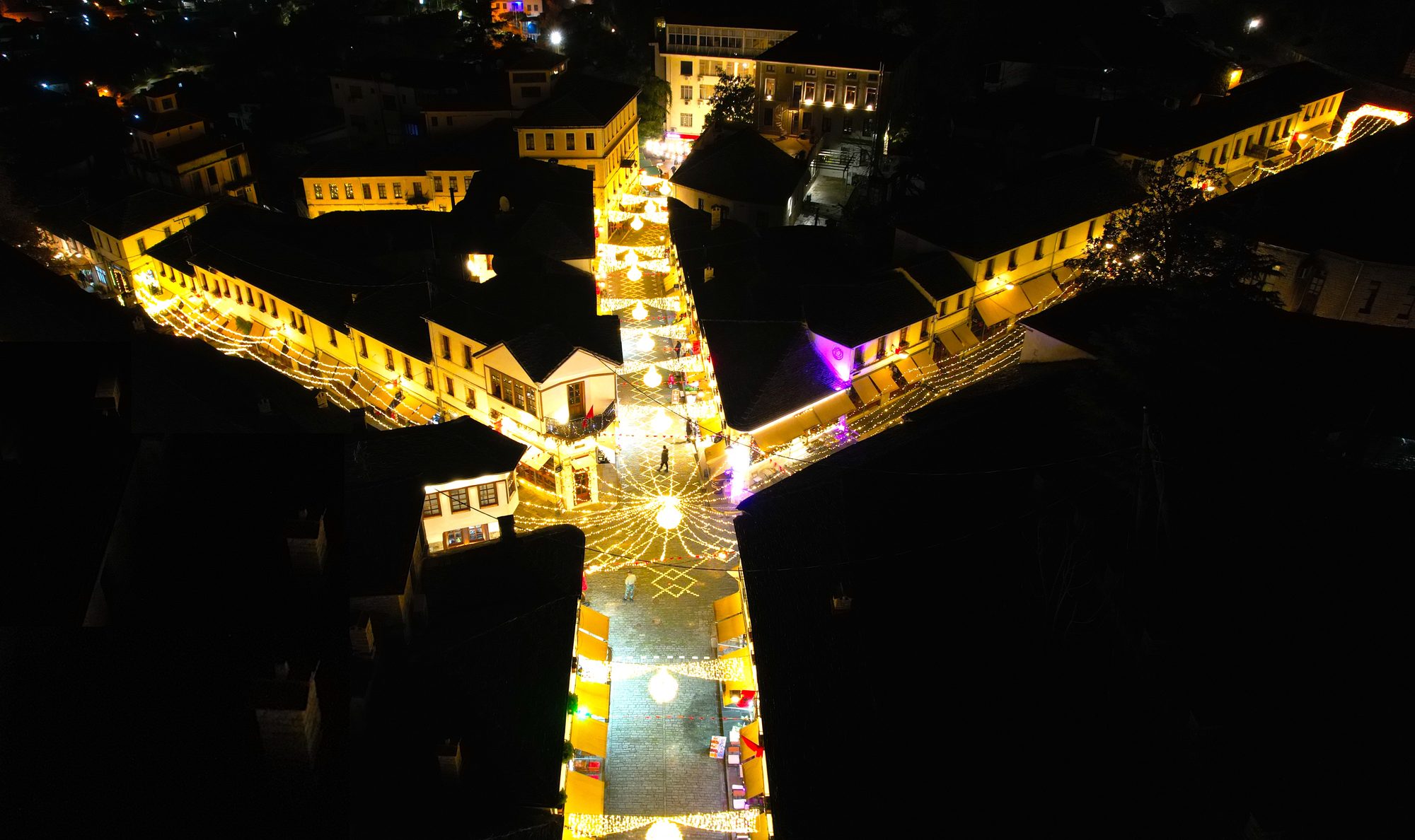 Aerial night view of Gjirokastër's old bazaar, its lanes strung with lights converging on the five-way junction, with dark slate roofs all around.