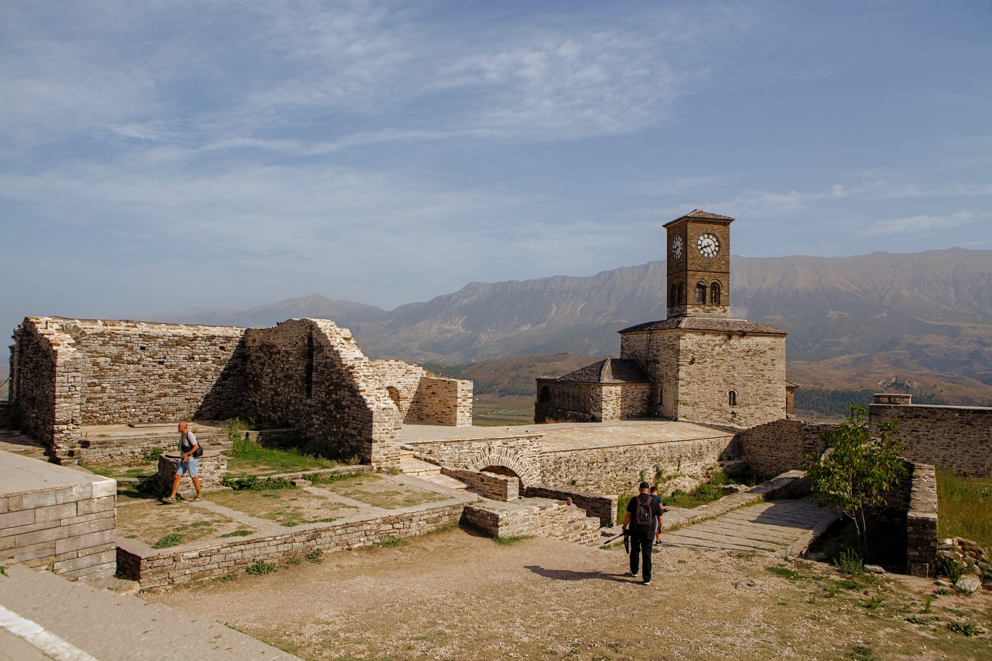 The stone clock tower inside Gjirokastër castle, rising above ruined walls and paved terraces, with visitors walking across and the Drino valley and mountains beyond.
