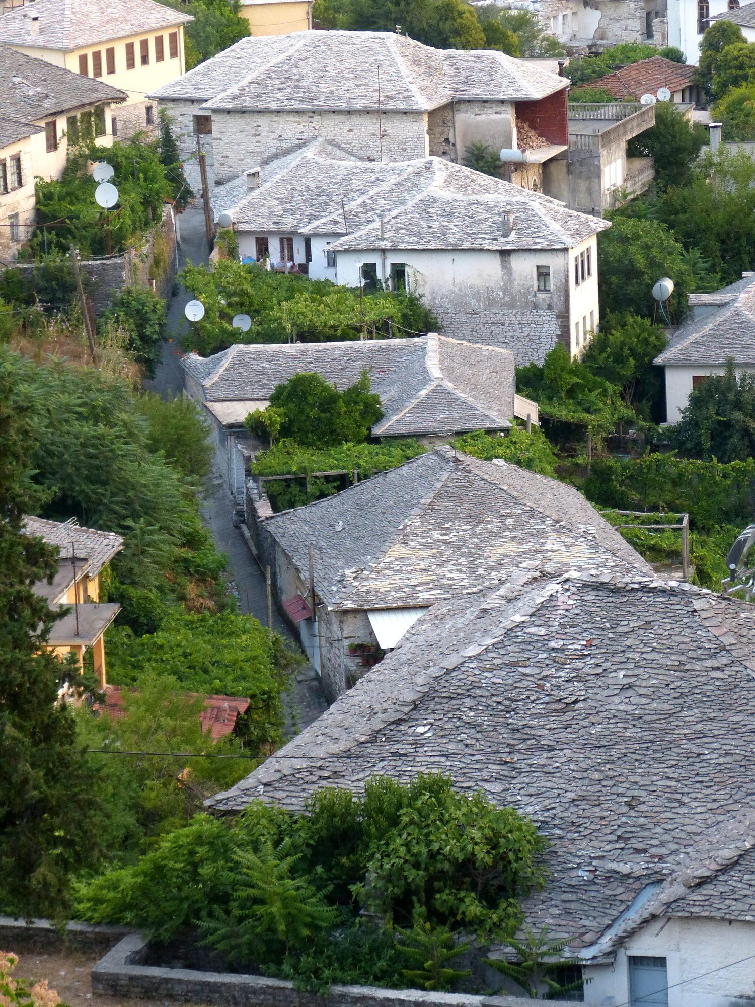 Steep stone-roofed Ottoman houses stacked on a hillside beneath a large castle, mountains behind.