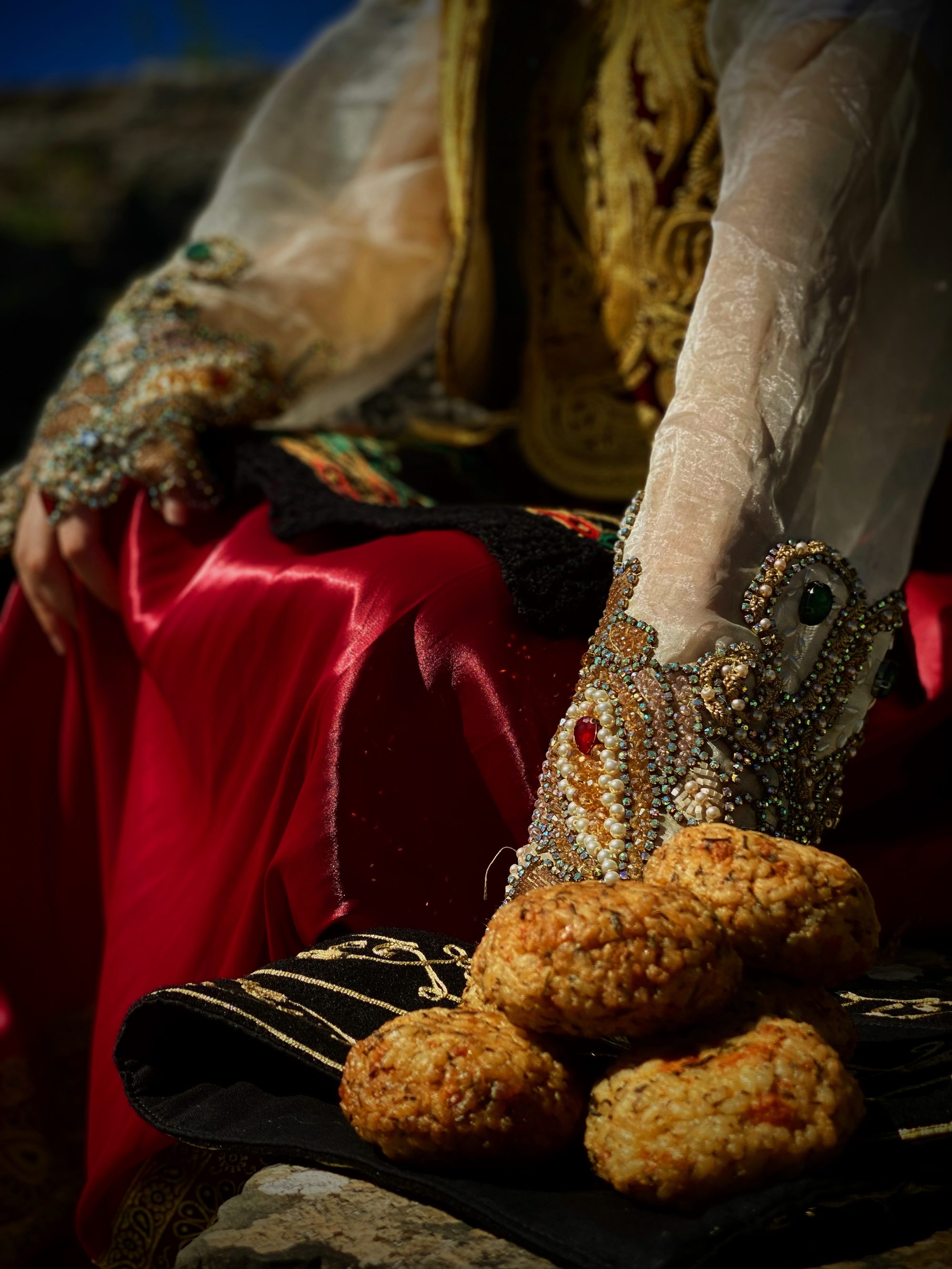 A pile of golden fried rice balls, qifqi, photographed against the red satin and gold embroidery of a traditional Gjirokastër costume.