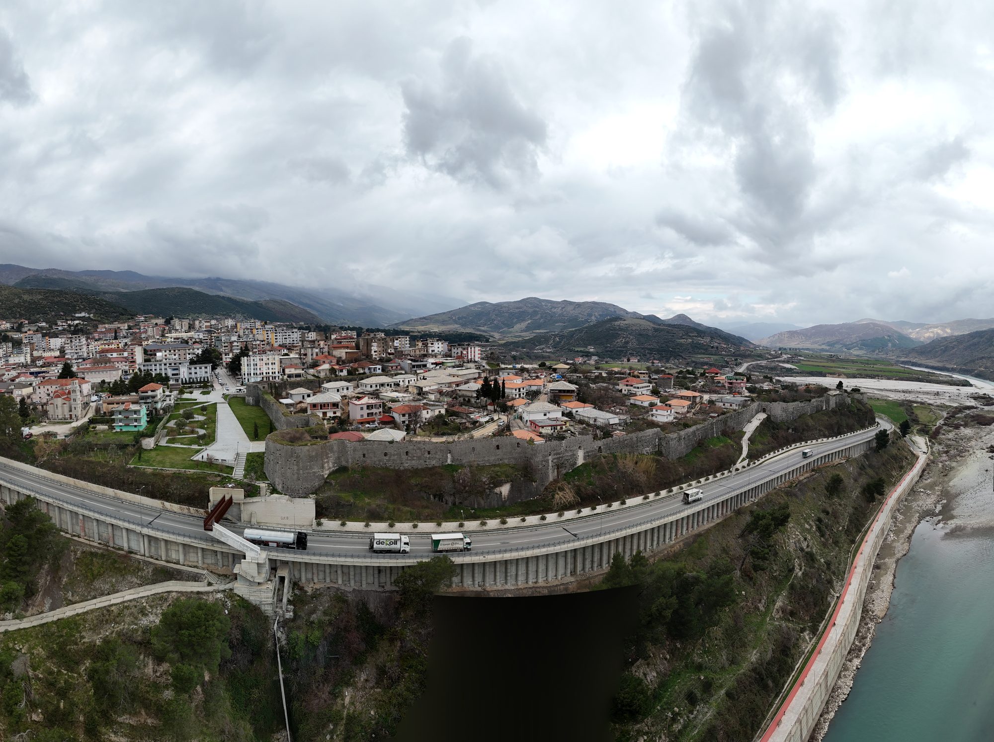 Aerial view of Tepelenë's castle walls enclosing a neighbourhood of houses, with the main road bridge crossing the Vjosa river in front and mountains behind.