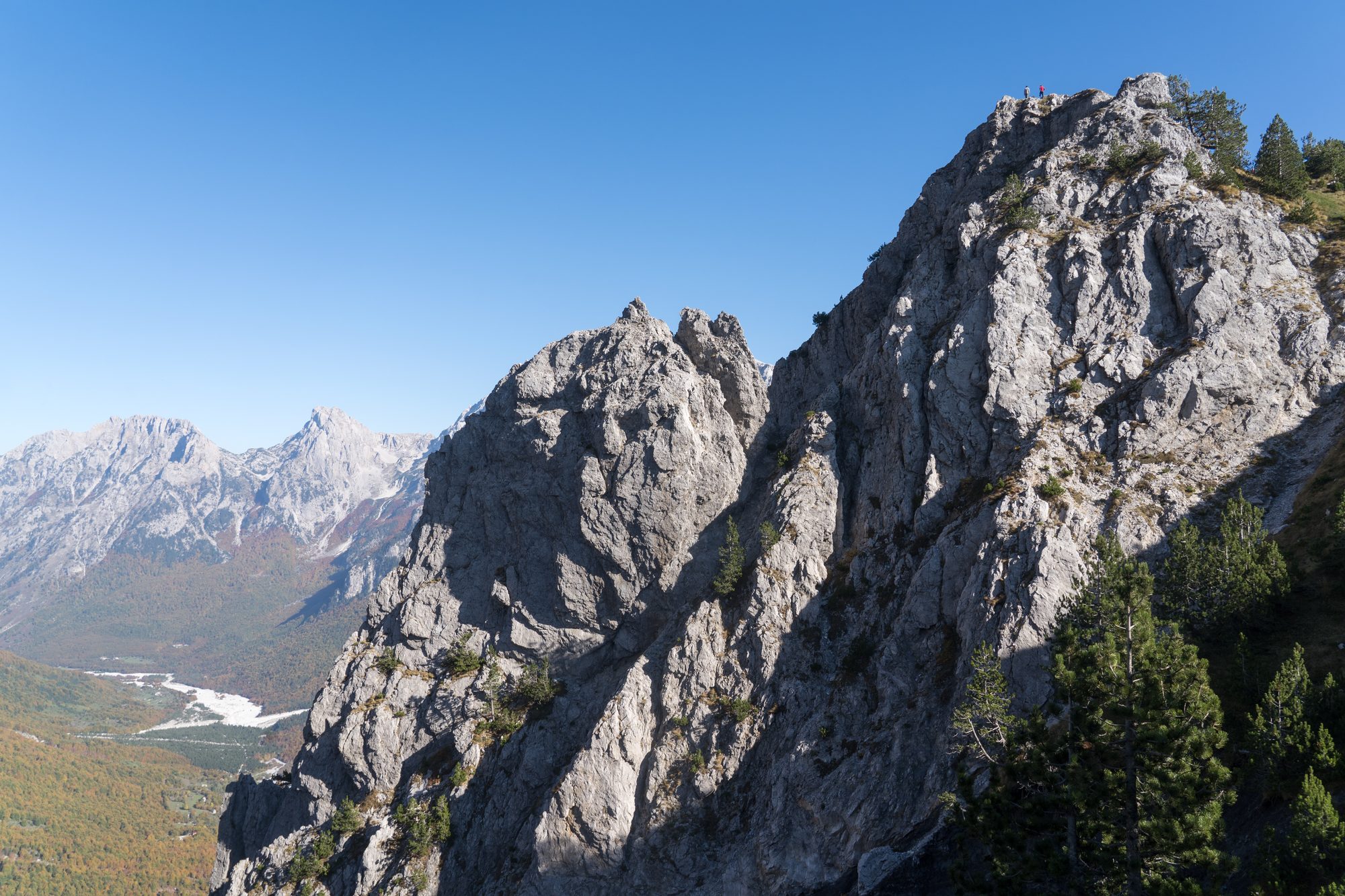 A jagged grey limestone ridge in the Valbona valley, with snow-flecked peaks behind it and the pale river bed far below.