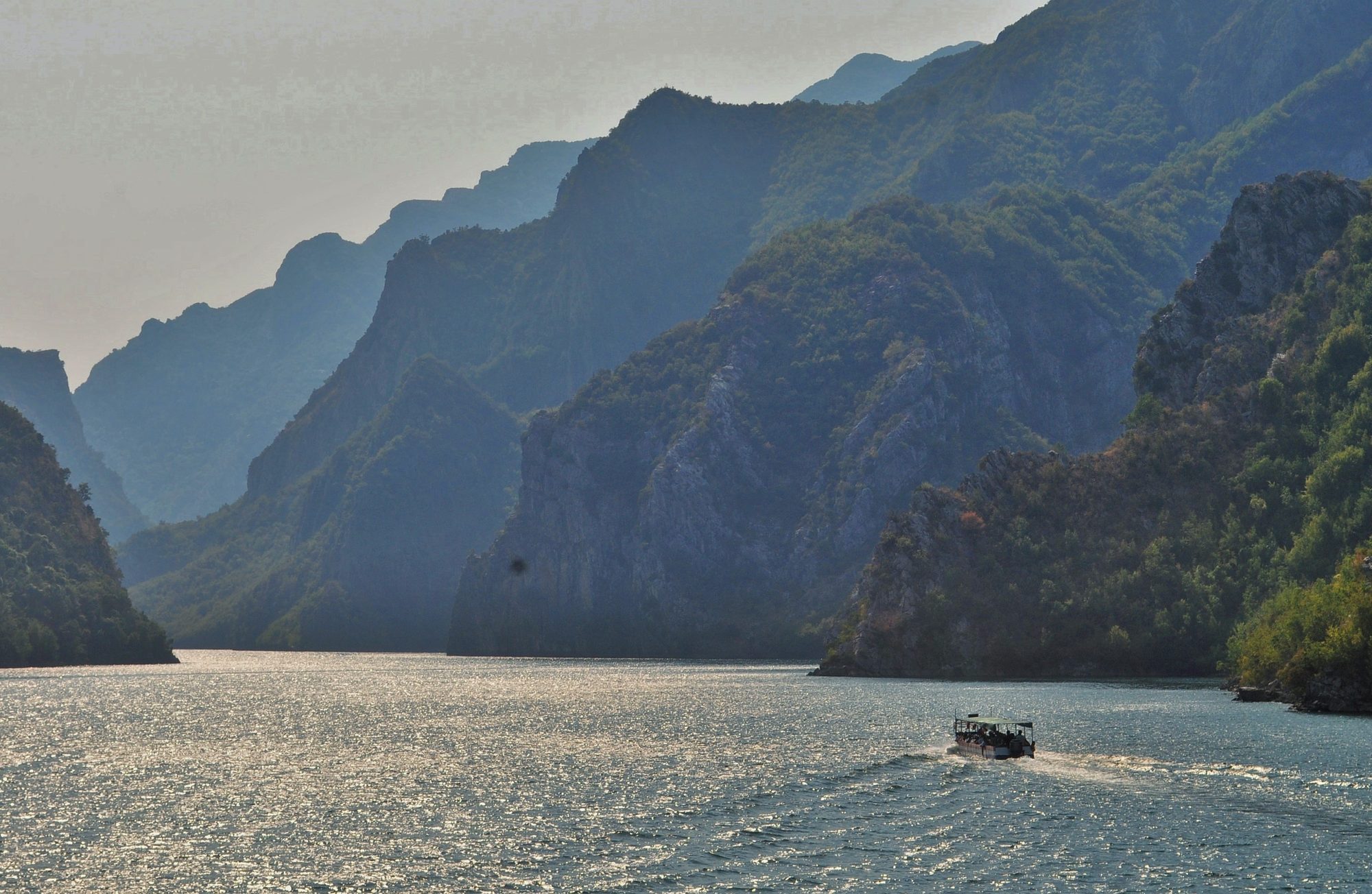 The steep canyon walls of Lake Koman rising straight out of still green water, seen from the ferry.