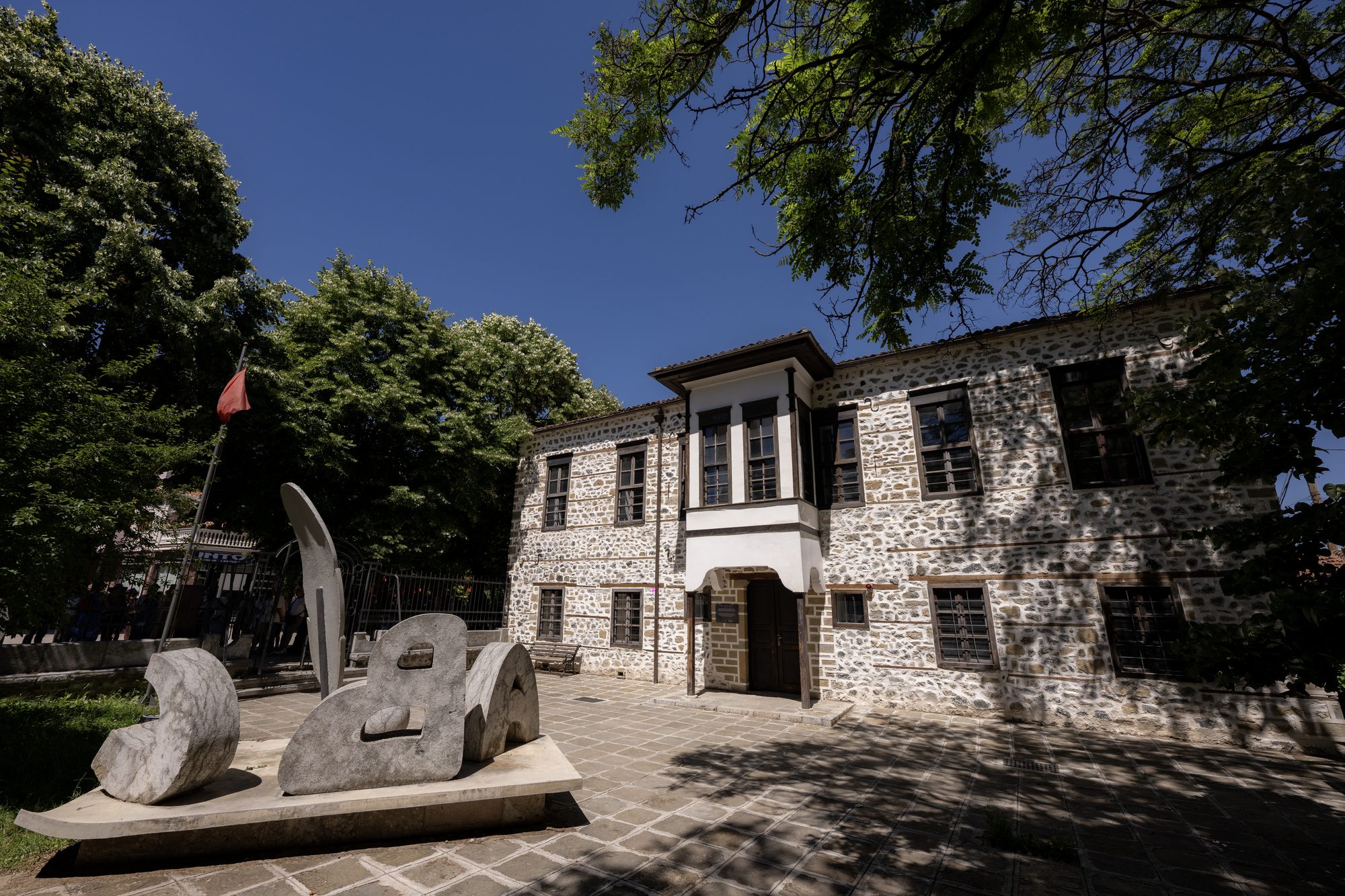 The two-storey stone building of the first Albanian-language school in Korçë, with a projecting timber bay window, a red flag and a stone sculpture of letters in the paved forecourt.