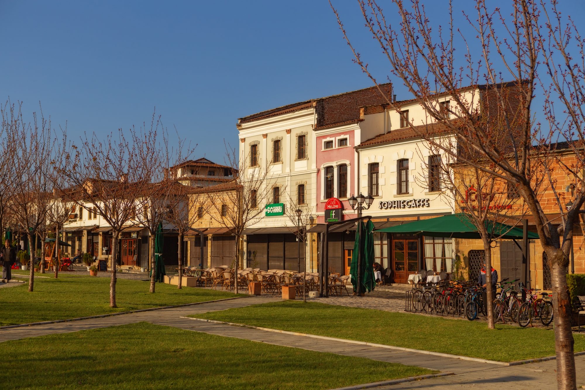 Restored two-storey Ottoman shopfronts along the edge of Korçë's Old Bazaar in early spring, with cafe terraces, parked bicycles and bare trees over a strip of lawn.