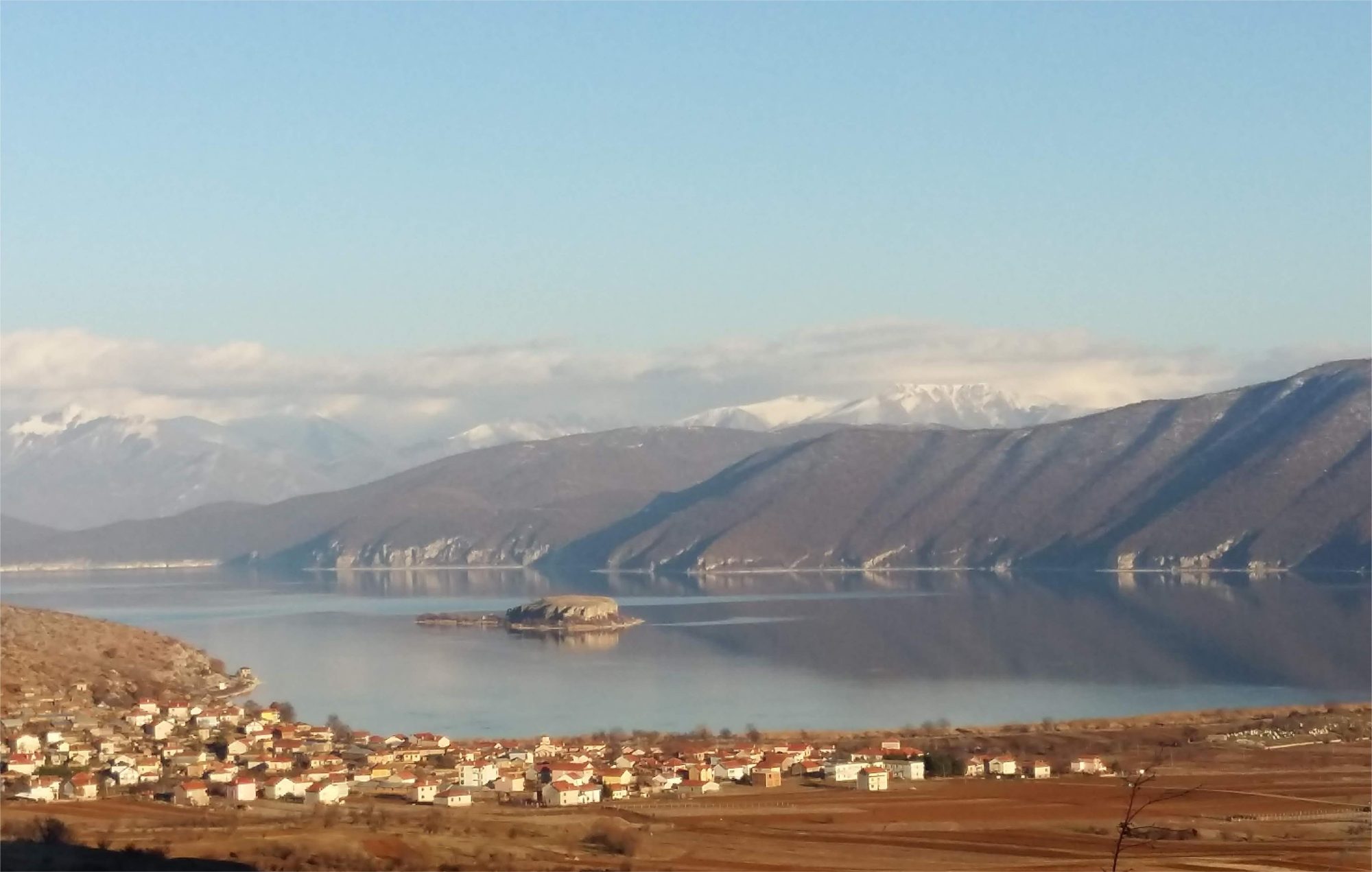 Great Prespa Lake seen from above Pustec, with a small rocky island in the bay, a village of red-roofed houses on the shore and snow-covered mountains beyond.
