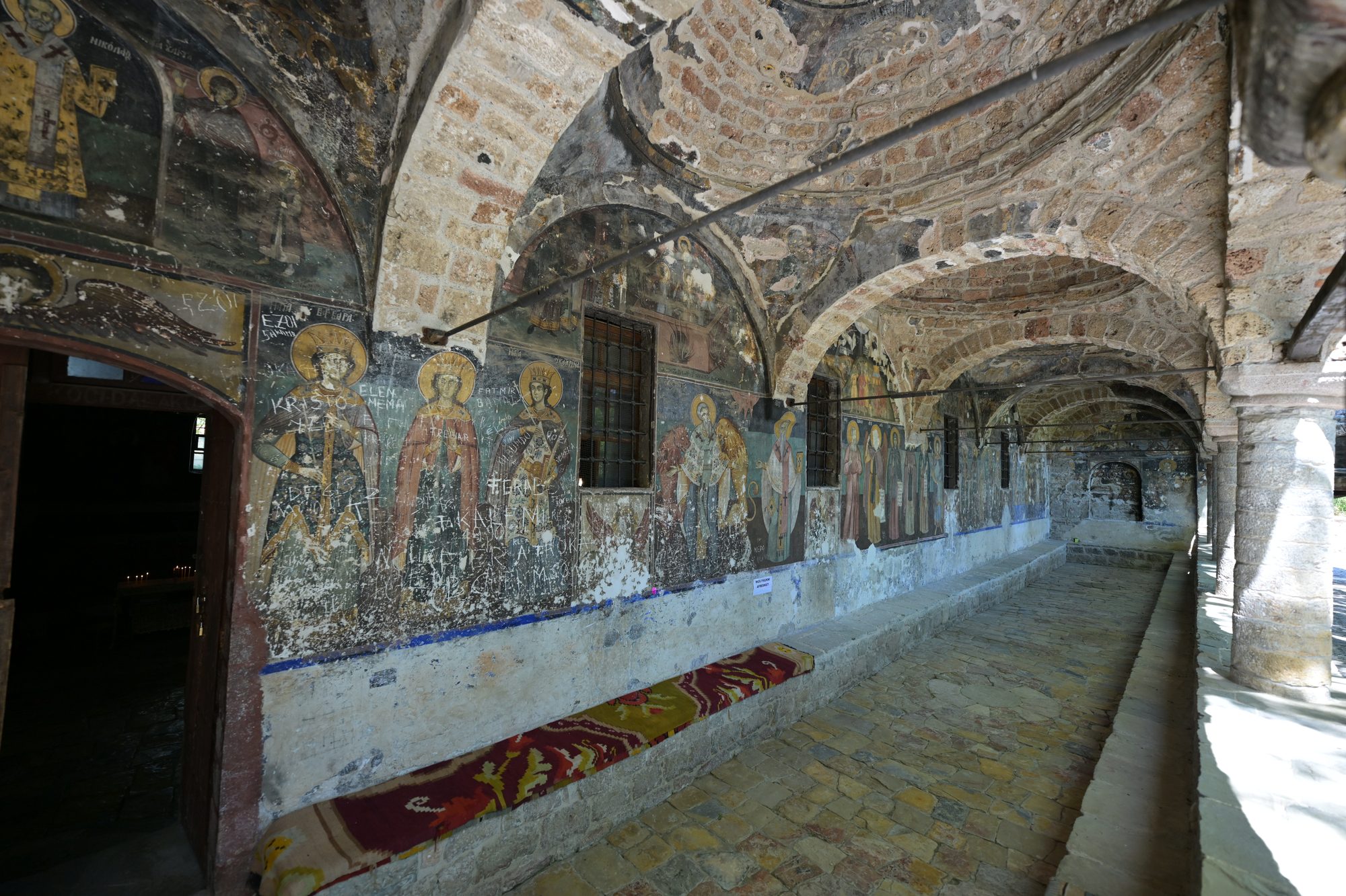 The frescoed open arcade of St Nicholas church in Voskopojë, with rows of painted saints under brick vaults and old graffiti scratched into the plaster.