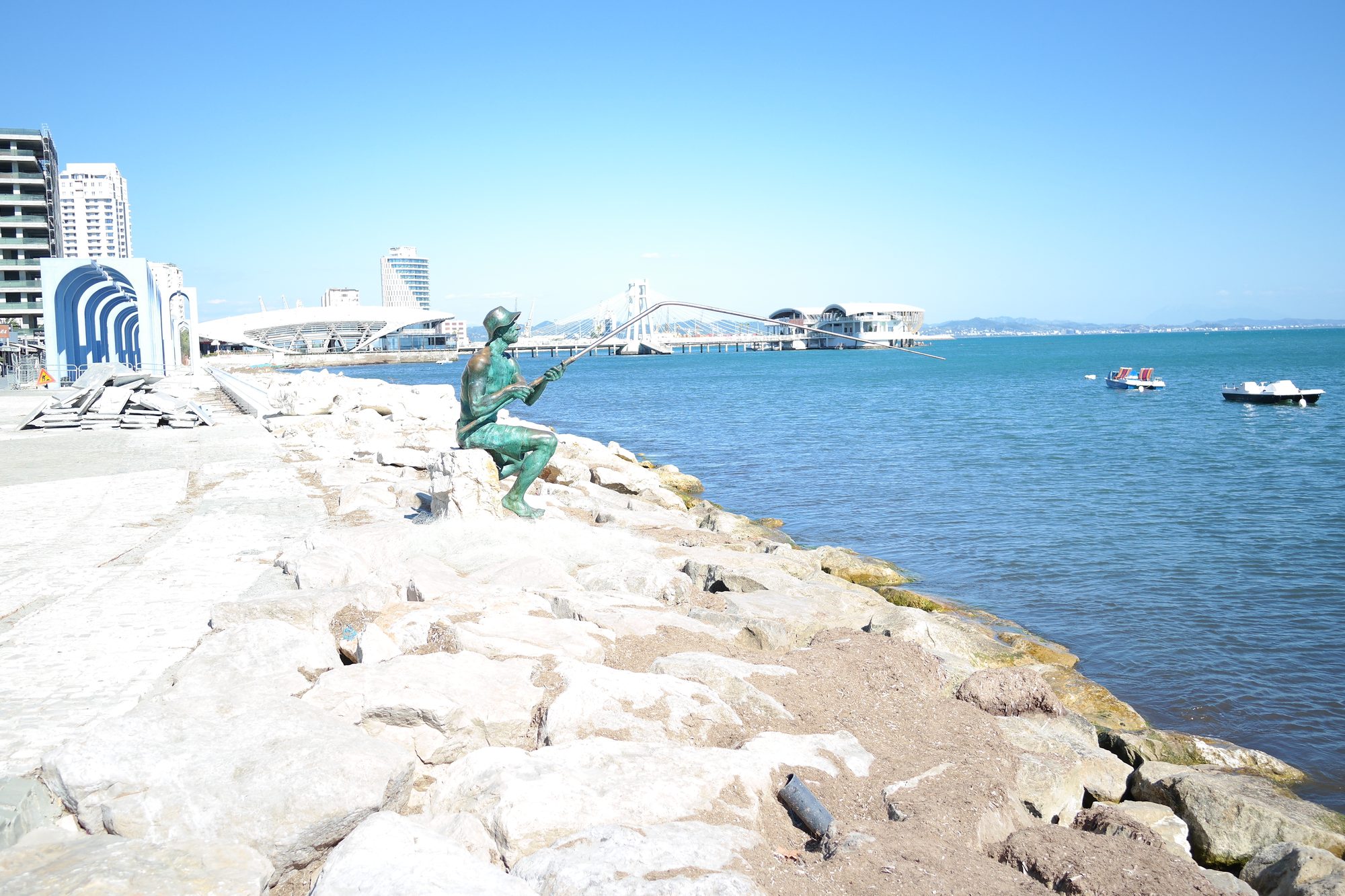 Bronze statue of a seated fisherman holding a rod on white rocks at the Durrës waterfront, with boats, a modern pier and the city in the background.