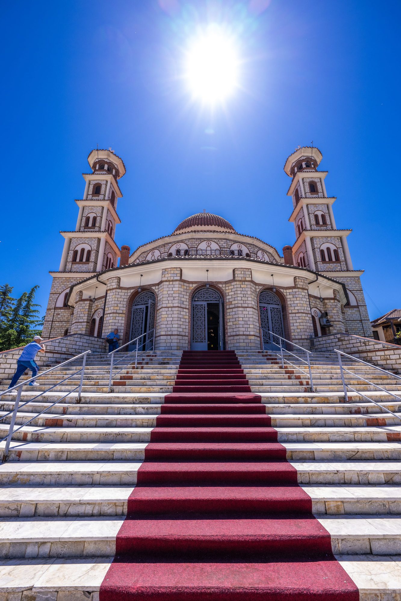 Front steps of the Resurrection Cathedral in Korçë, a red carpet running up white stone stairs toward twin bell towers and a dome under a deep blue sky.