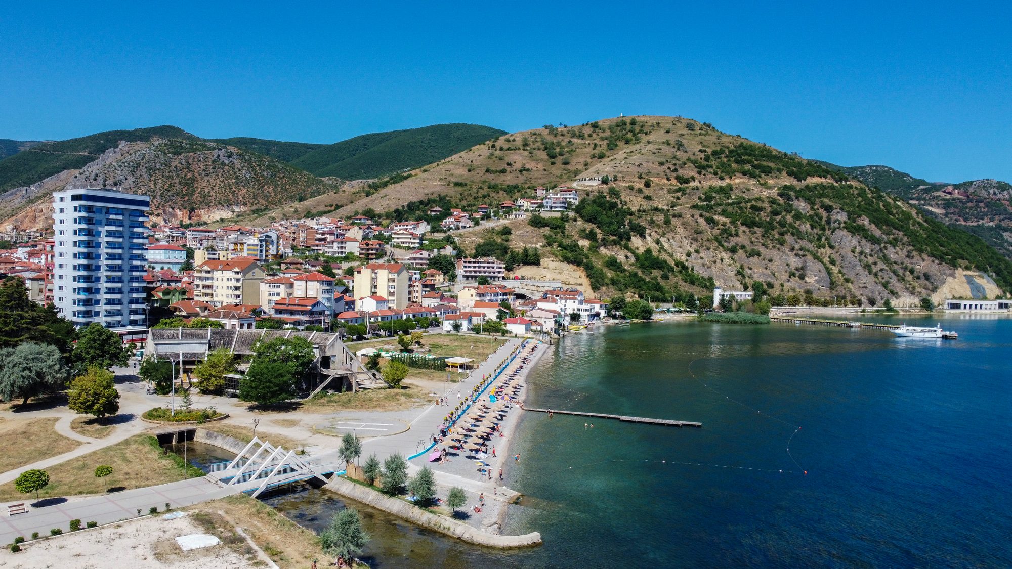 Aerial view of Pogradec's shoreline in summer, with a town beach full of umbrellas and swimmers, a jetty, apartment blocks and dry mountains behind.
