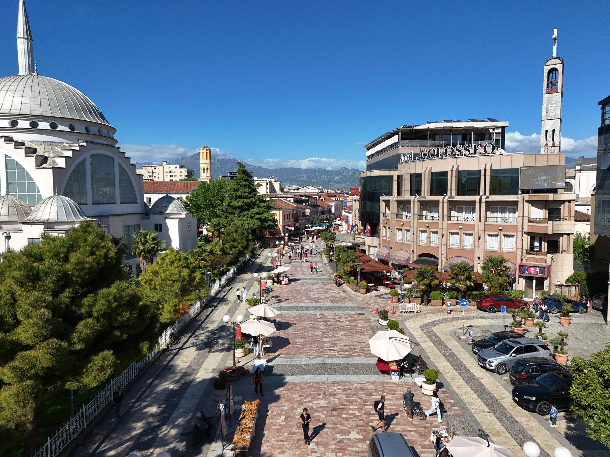Aerial view of Shkodra's pedestrian Kolë Idromeno street with cafe umbrellas and stalls, the dome of a mosque on the left and a clock tower behind.