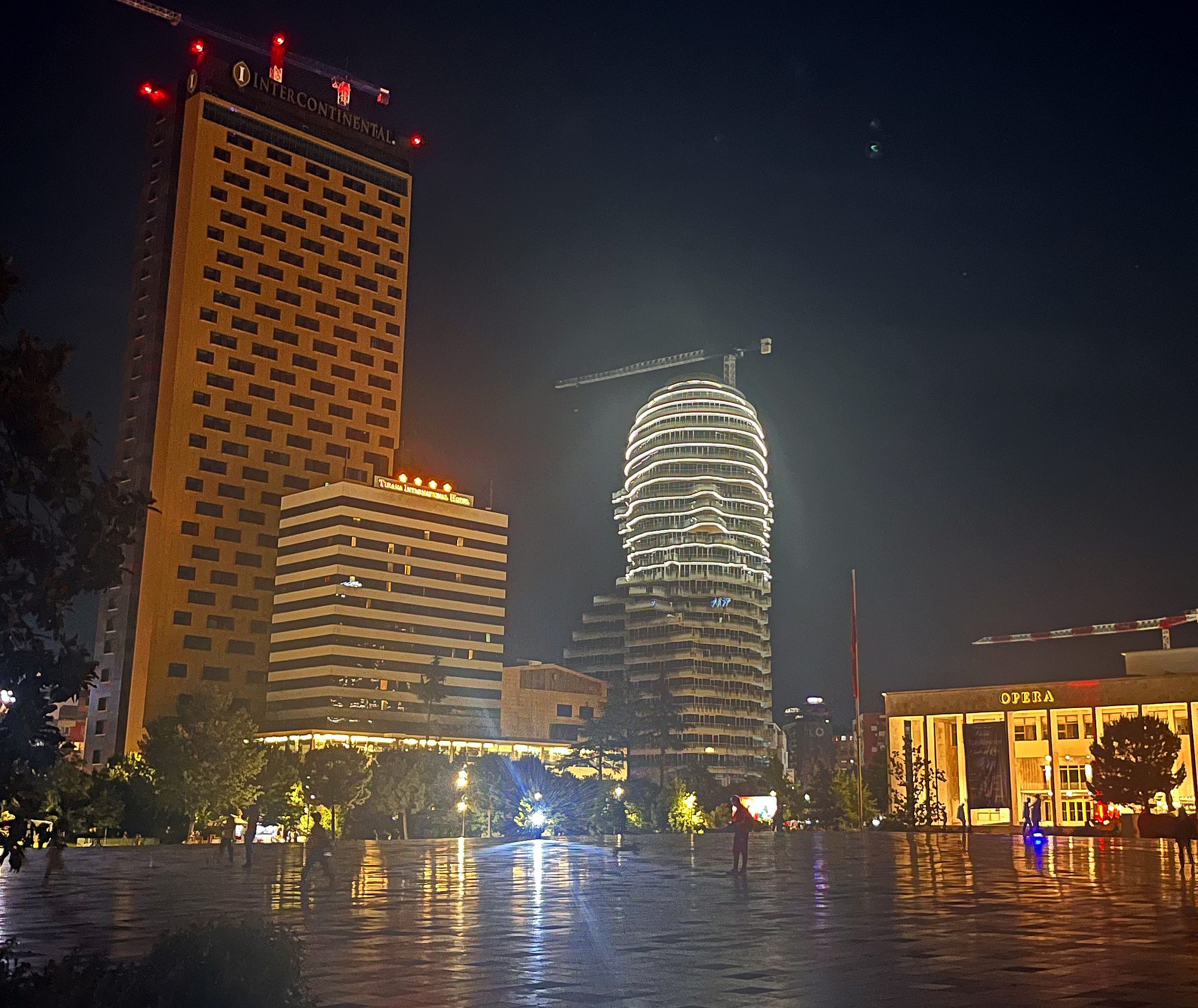 Skanderbeg Square in Tirana at night, wet paving reflecting the lights of the Opera, the Tirana International Hotel and an illuminated tower.