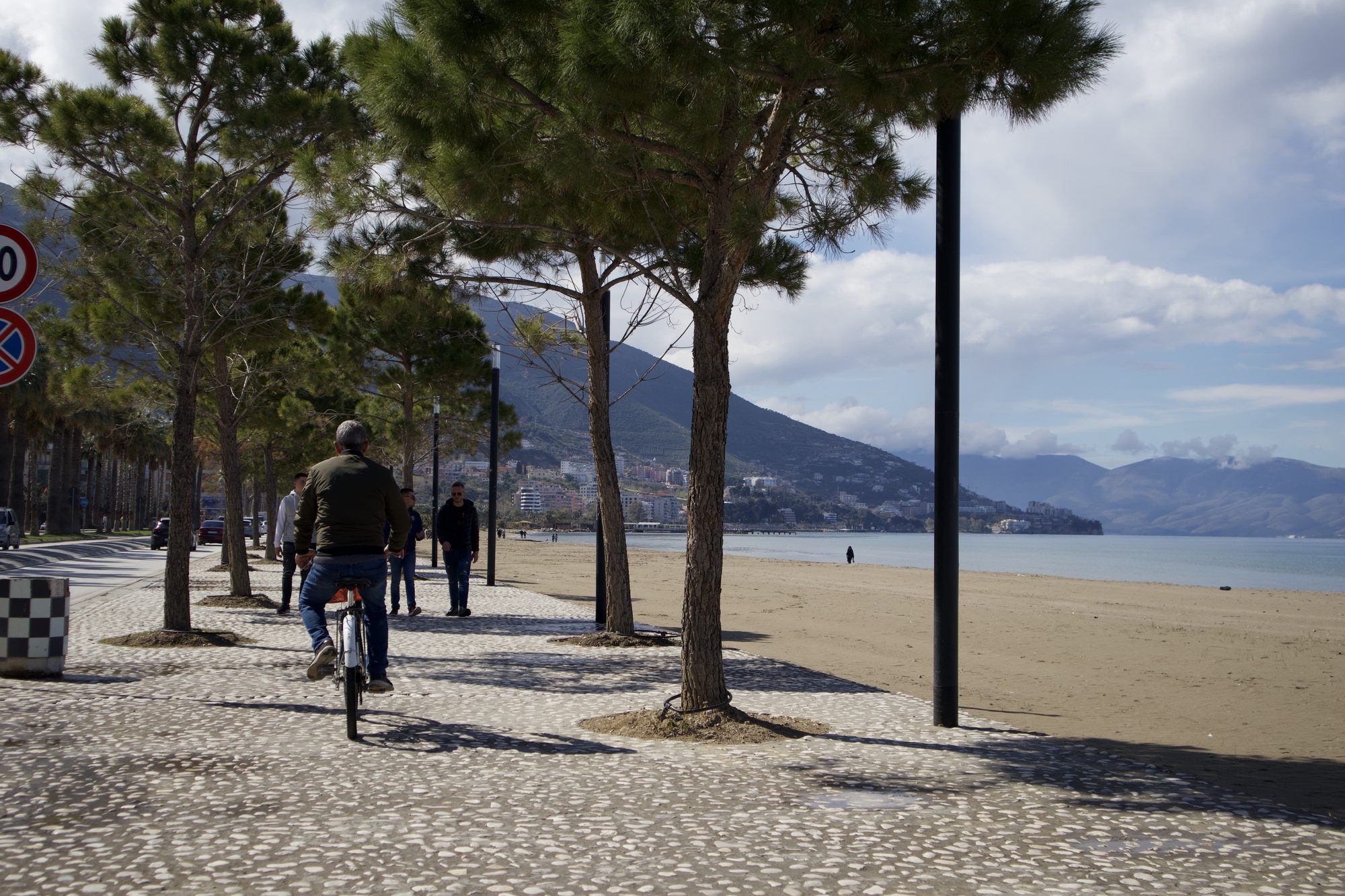 Mosaic-paved promenade in Vlorë lined with pine trees, people strolling and a man cycling, with the town beach and the bay alongside.