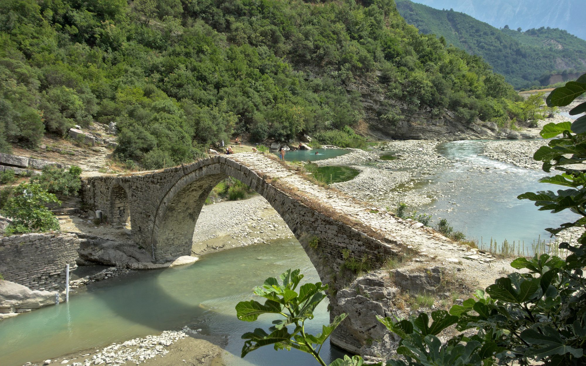 Ottoman stone bridge arching over a river beside natural thermal pools where a few people are bathing, wooded hills behind.
