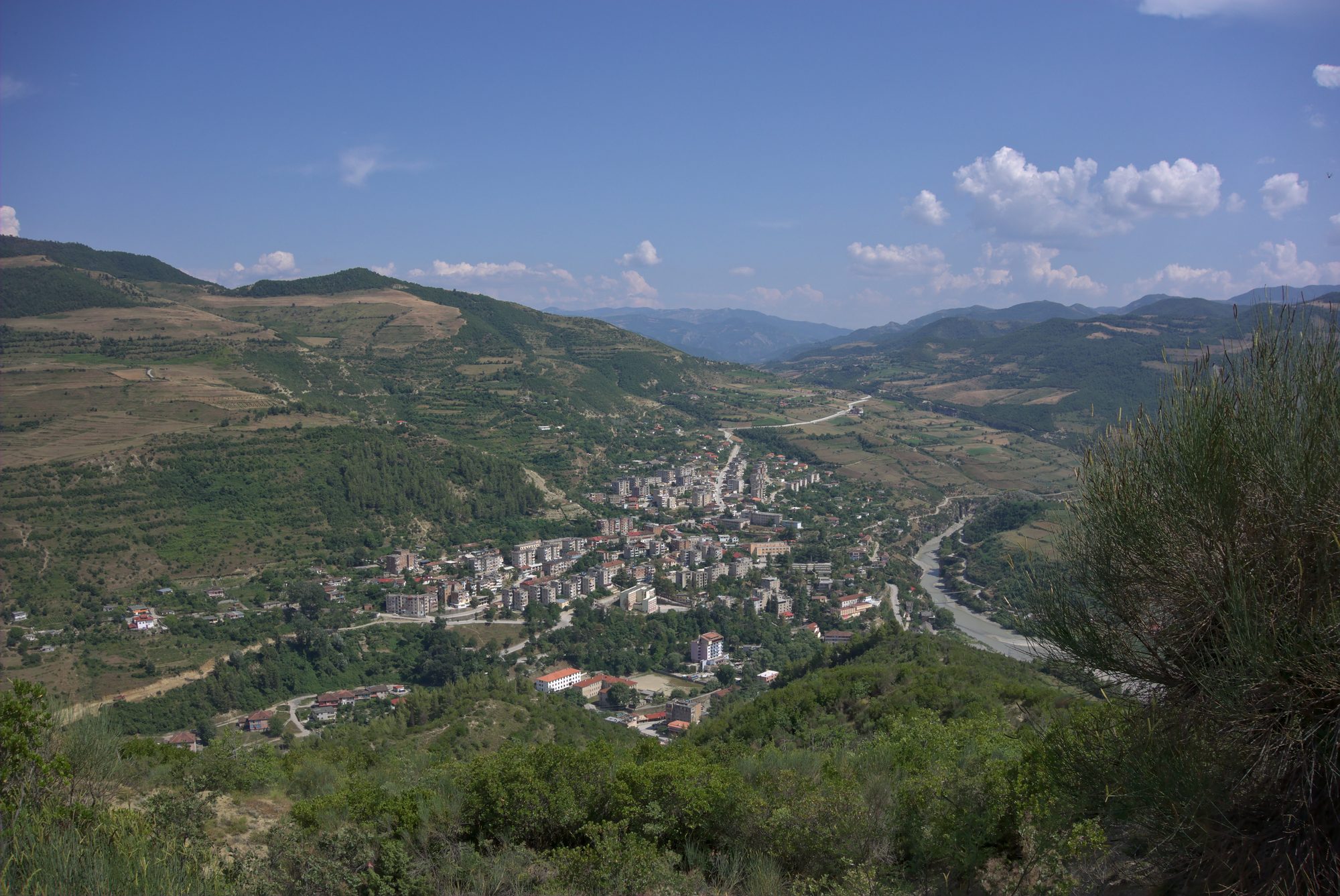 Çorovoda seen from a hillside above, apartment blocks and houses along a river valley surrounded by mountains.