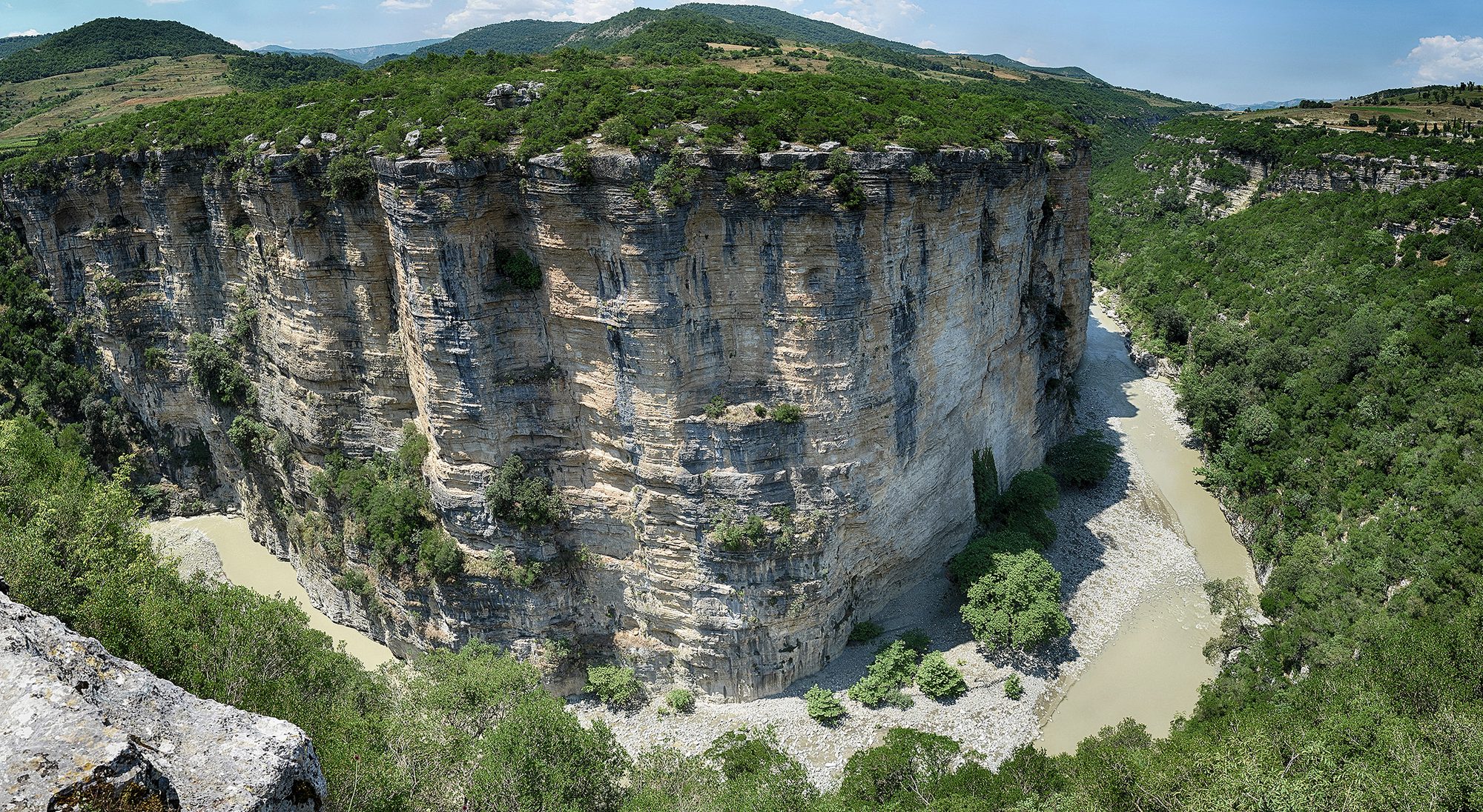 Limestone walls of the Osumi canyon curving around a bend of the river, seen from the rim with green hills behind.