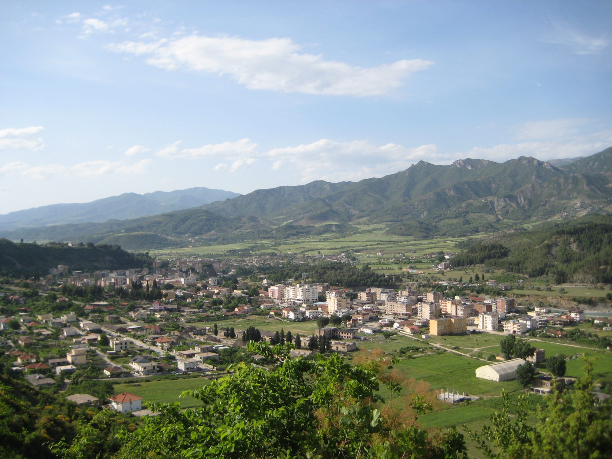 Përmet town in a green river valley with apartment buildings and houses, high mountains rising behind under a blue sky.