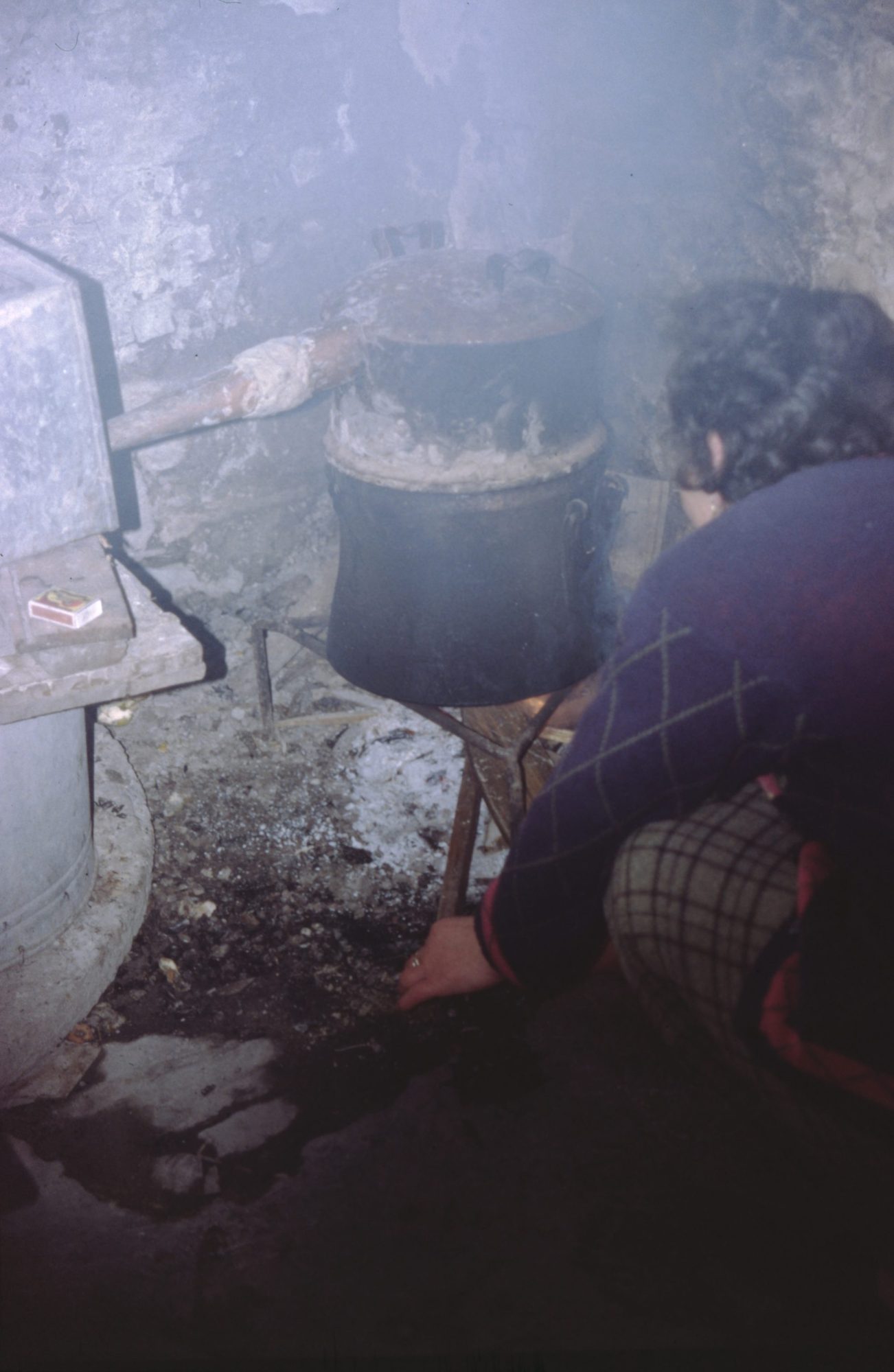 A woman crouches beside a wood fire under a blackened pot still in a smoky stone room, tending the embers with her hand.