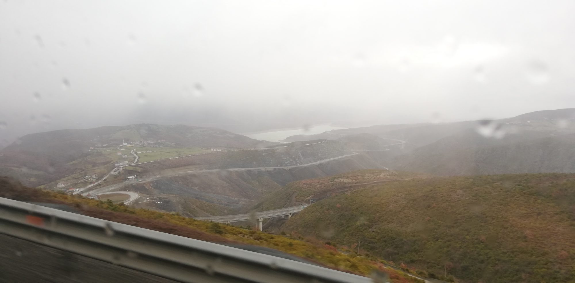 A wet mountain road in heavy rain, with low cloud obscuring the slopes and poor visibility ahead.