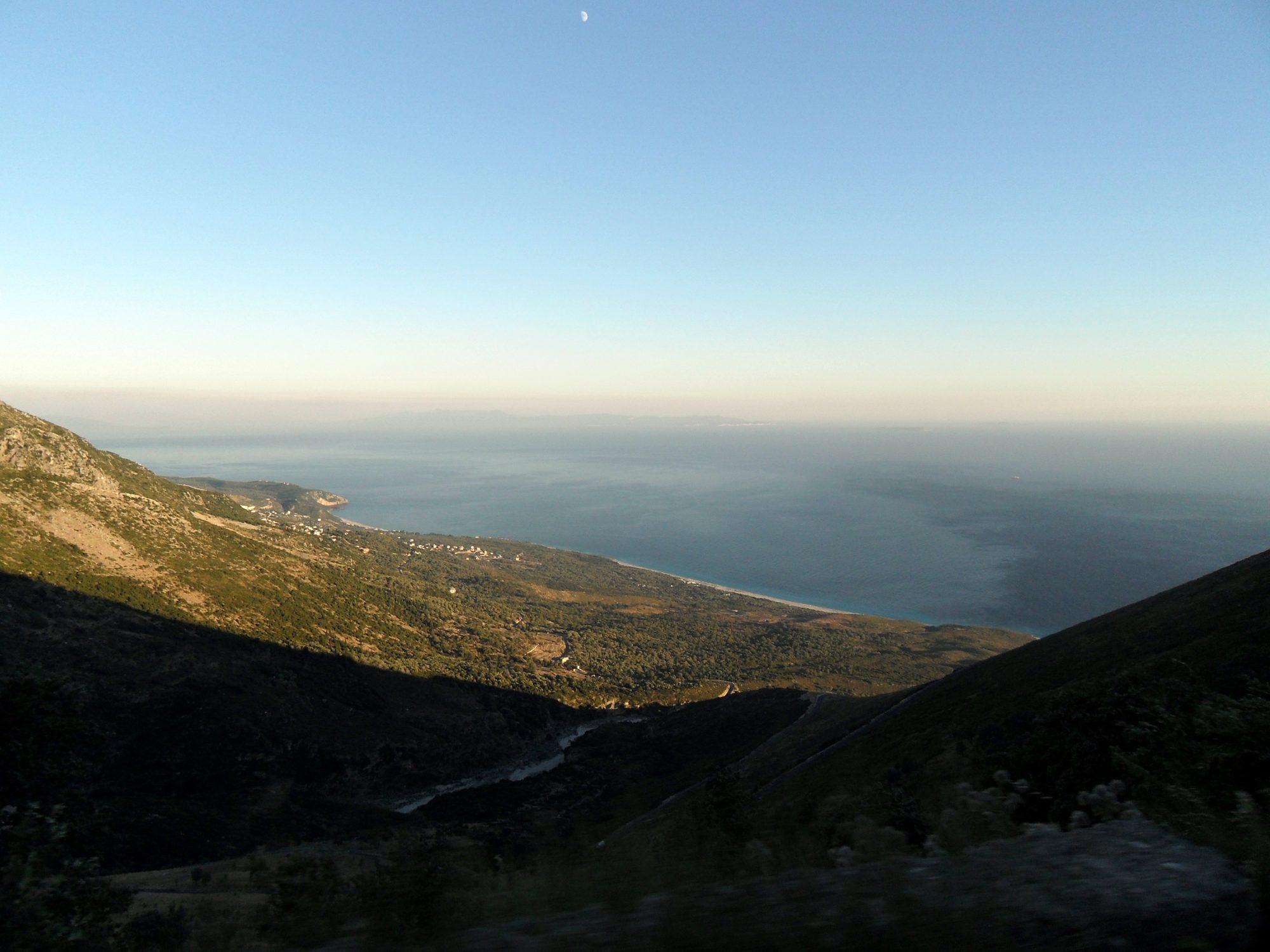 A narrow asphalt road curving along a steep hillside above the sea, with no barrier on the drop side.