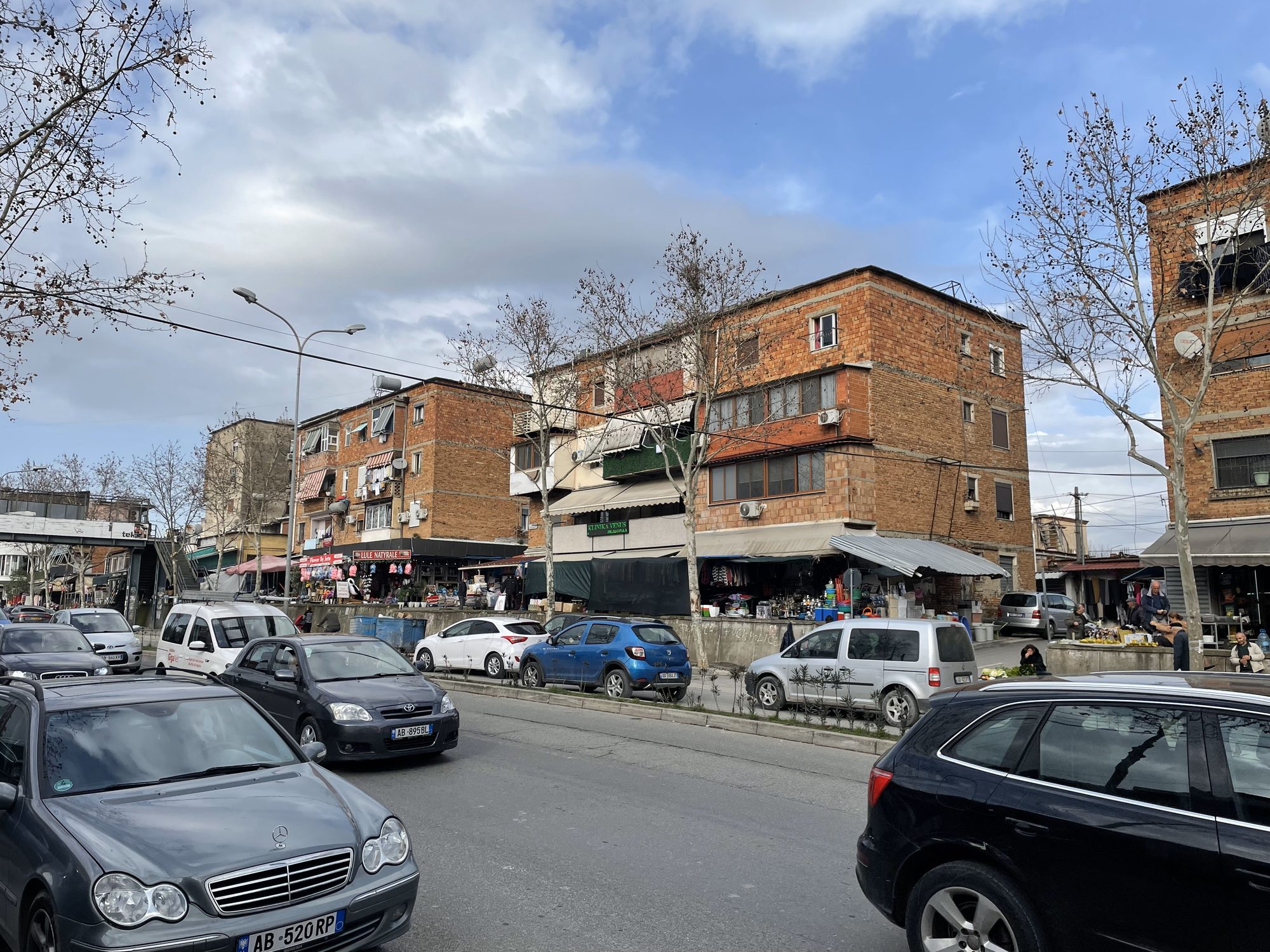 A busy road with cars in both directions and a roadside market with stalls and pedestrians close to the traffic.