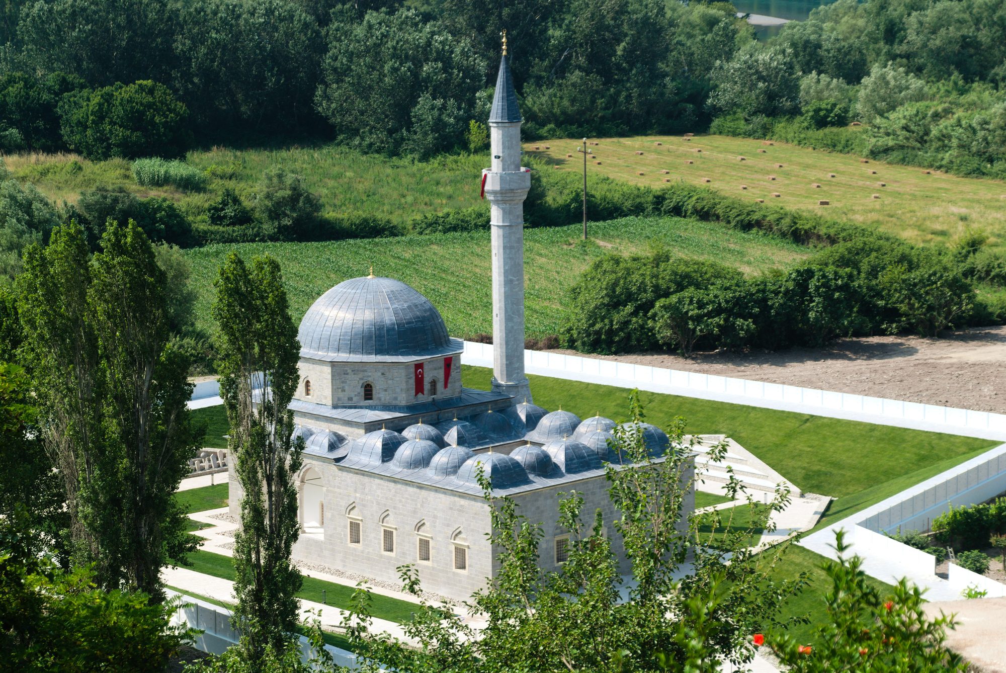 Stone mosque with a cluster of grey lead-covered domes and a single slender minaret standing among green fields and poplar trees.