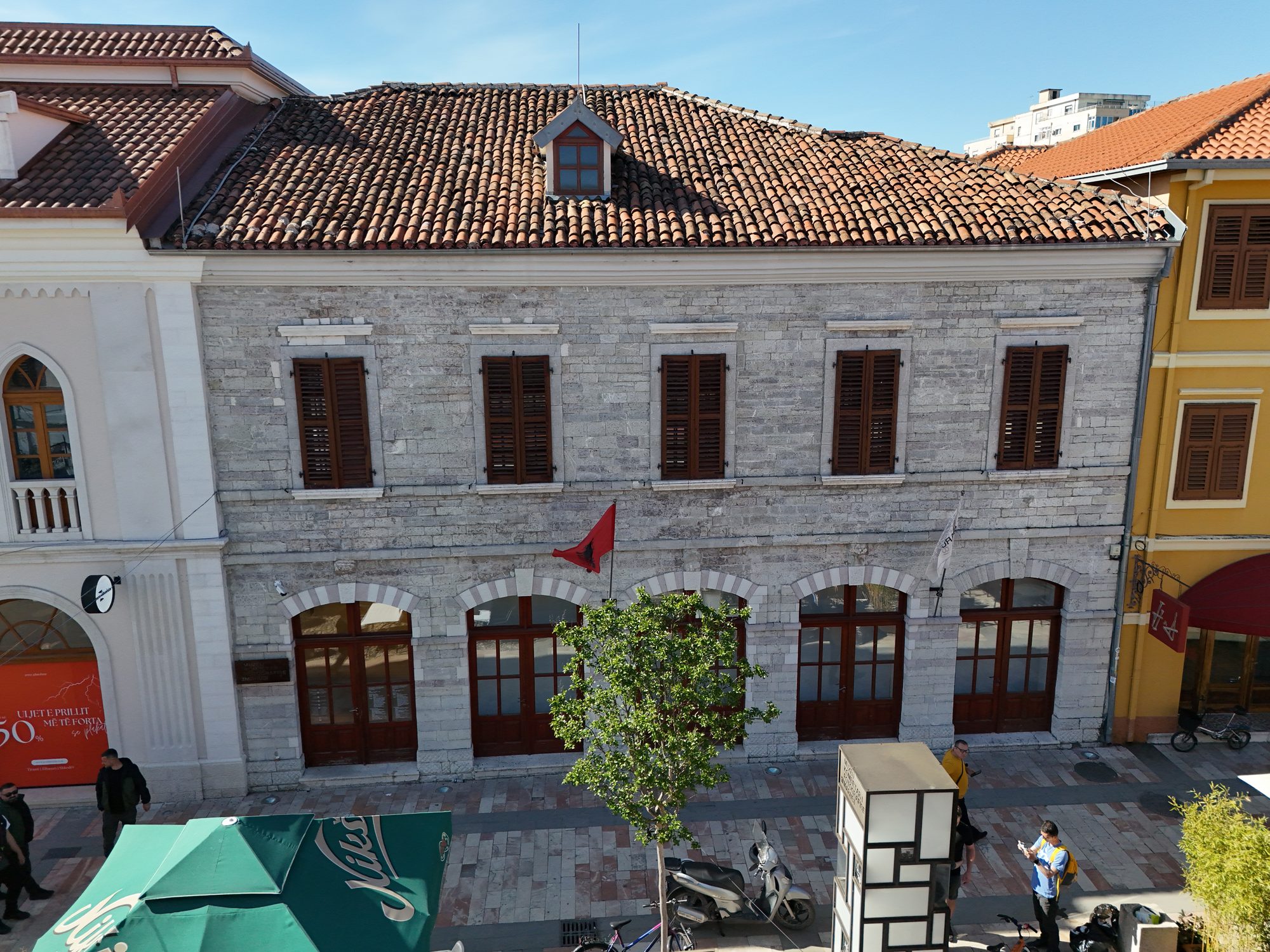 Two-storey stone building with tall arched windows and wooden shutters on a pedestrian street, an Albanian flag by the door and a cafe umbrella at the corner.