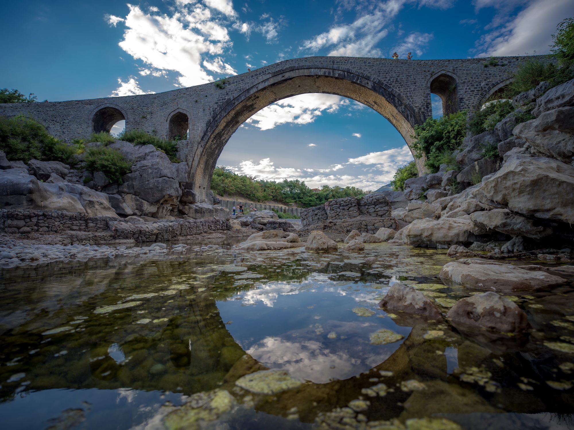 Tall stone arch bridge with several smaller arches spanning a rocky riverbed, its reflection visible in a still green pool below.