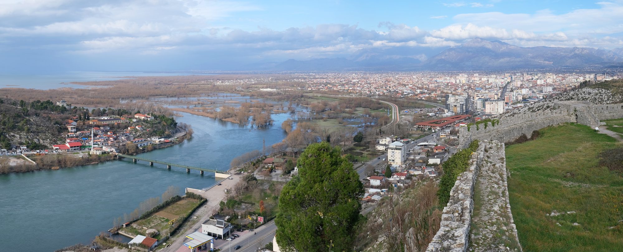 View from the stone walls of Rozafa Castle over the Buna river, a road bridge, the flat city of Shkodër and mountains on the horizon.