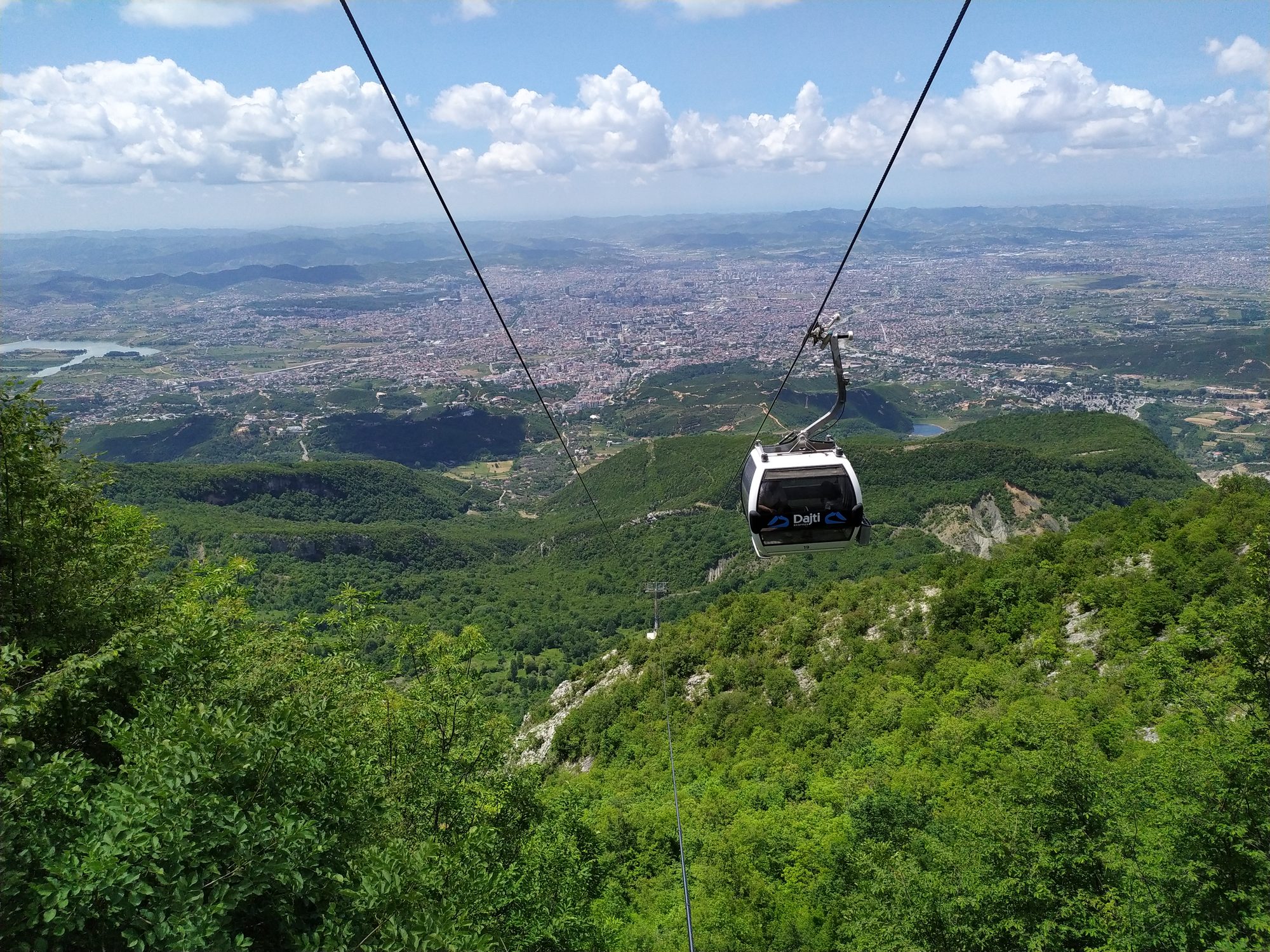 A red and white cable car cabin suspended above forested slopes, with hills receding into haze behind.