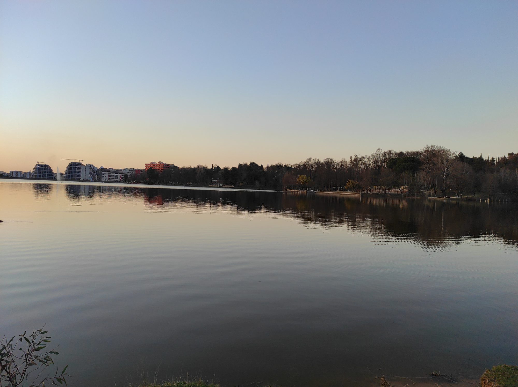 A tree-lined path beside a lake in a city park, with people walking in the late afternoon light.