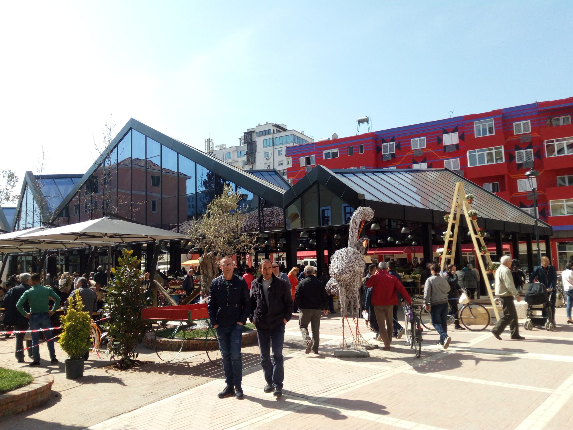 Stalls of fruit and vegetables under bright awnings in a paved market square surrounded by low buildings.